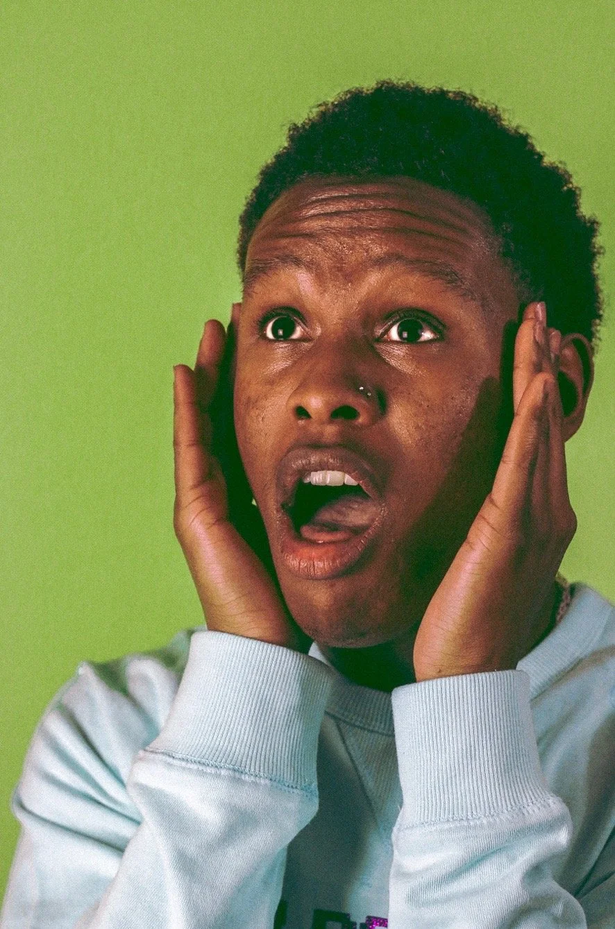 A woman with short curly hair and a nose piercing, expressing shock or surprise with her mouth open and hands on her face, against a green background.