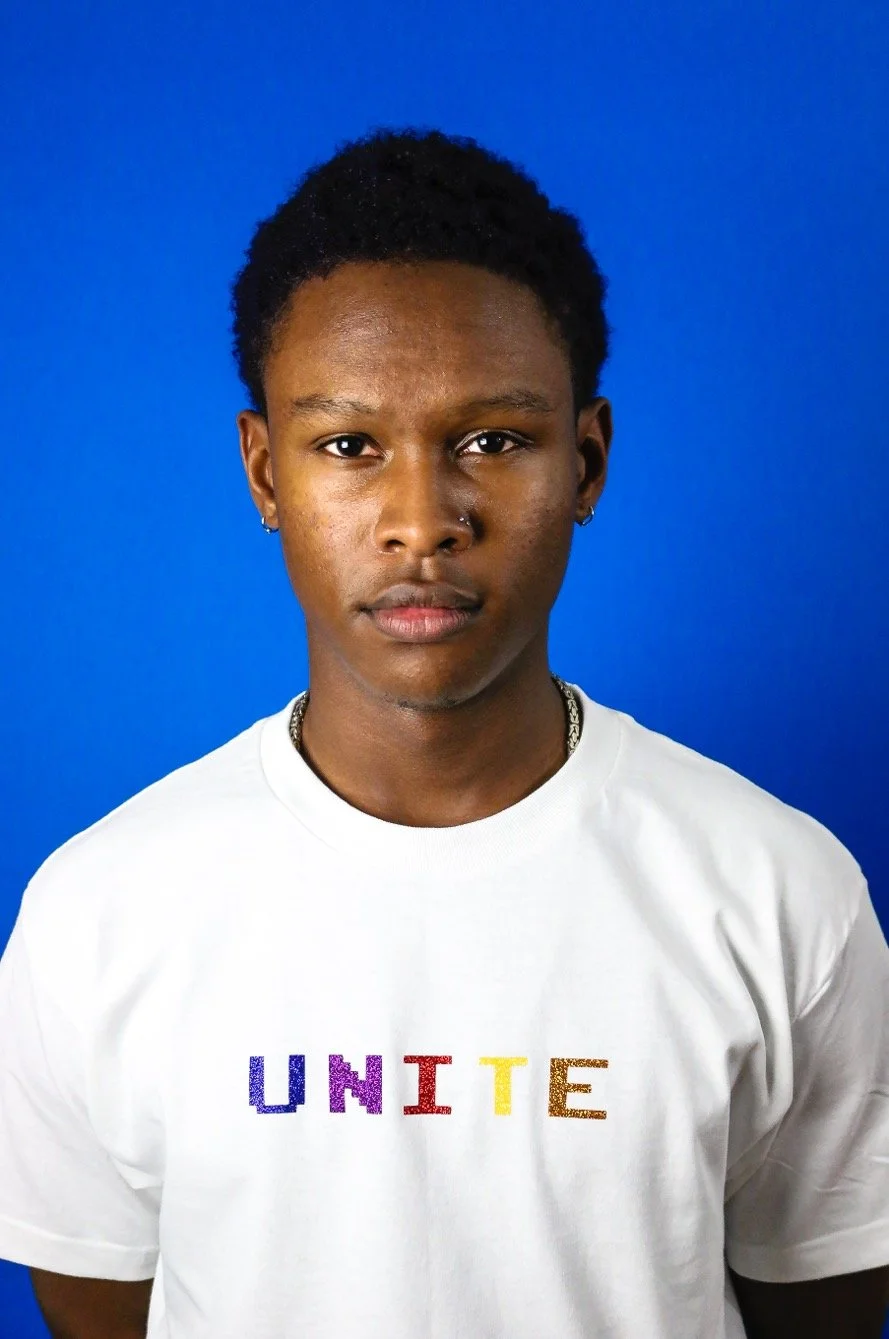 Young person with short curly hair and earrings wearing a white t-shirt with the word 'UNITE' in rainbow colors, standing against a blue background.
