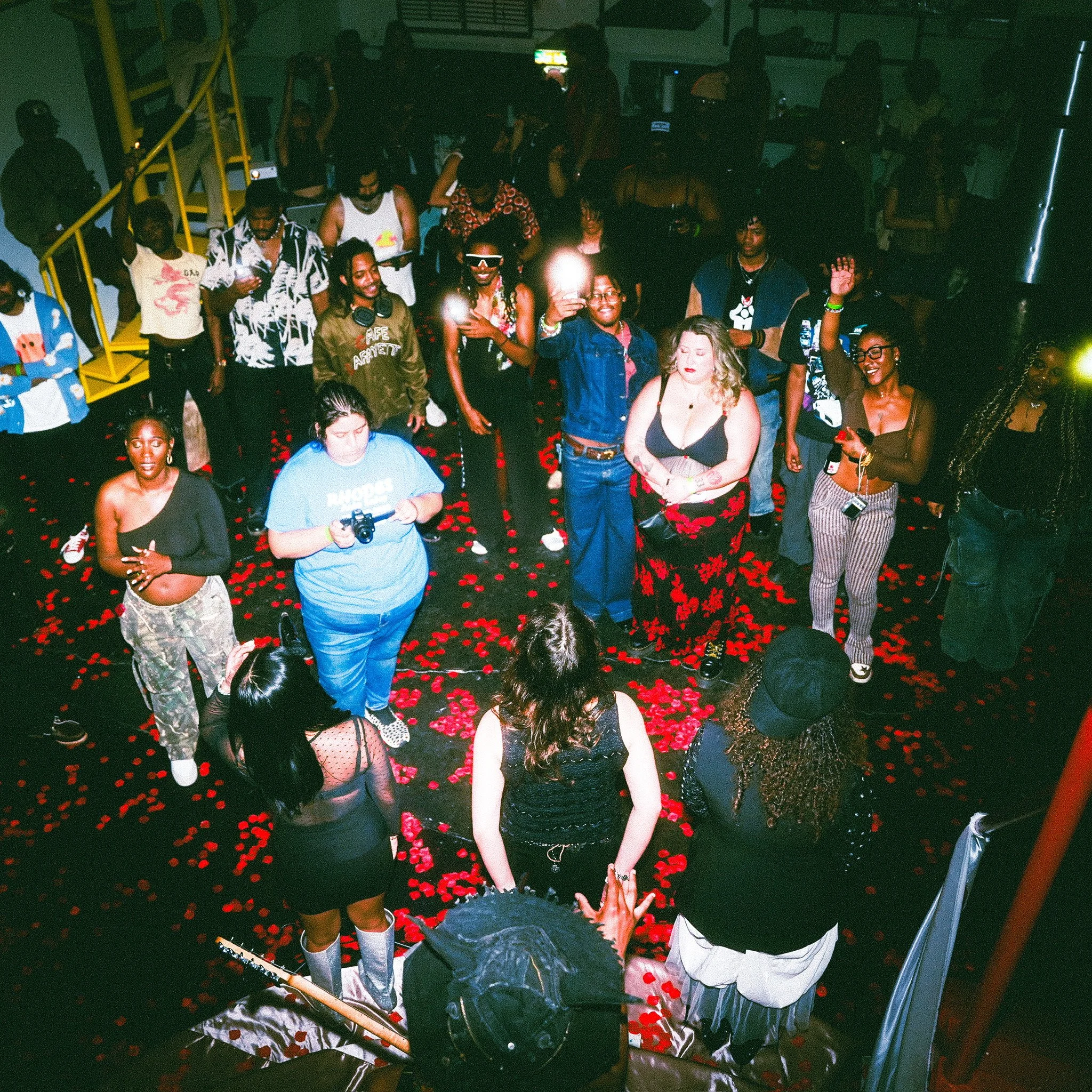 Group of people dancing and socializing on a dance floor covered with red flower petals, with some holding drinks and a person with a camera in the foreground, in a dimly lit indoor setting.