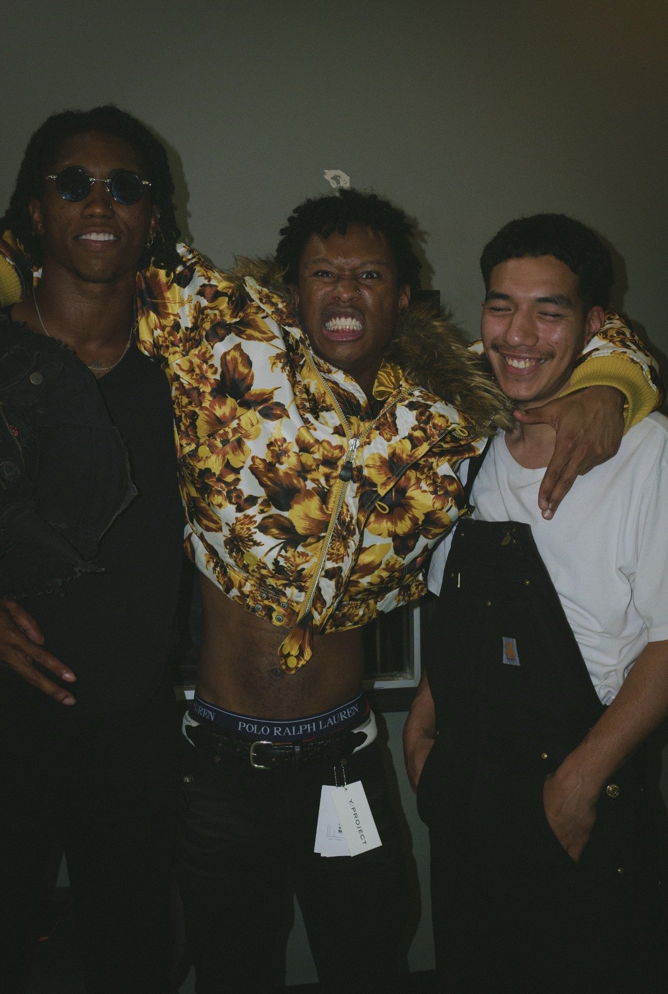 Three young men standing close together, smiling and showing friendship. One in the middle is shirtless, wearing a floral jacket and Polo Ralph Lauren underwear waistband. The man on the left wears sunglasses and a black jacket, while the man on the right wears a white shirt. They appear to be indoors, possibly at a party or social gathering.