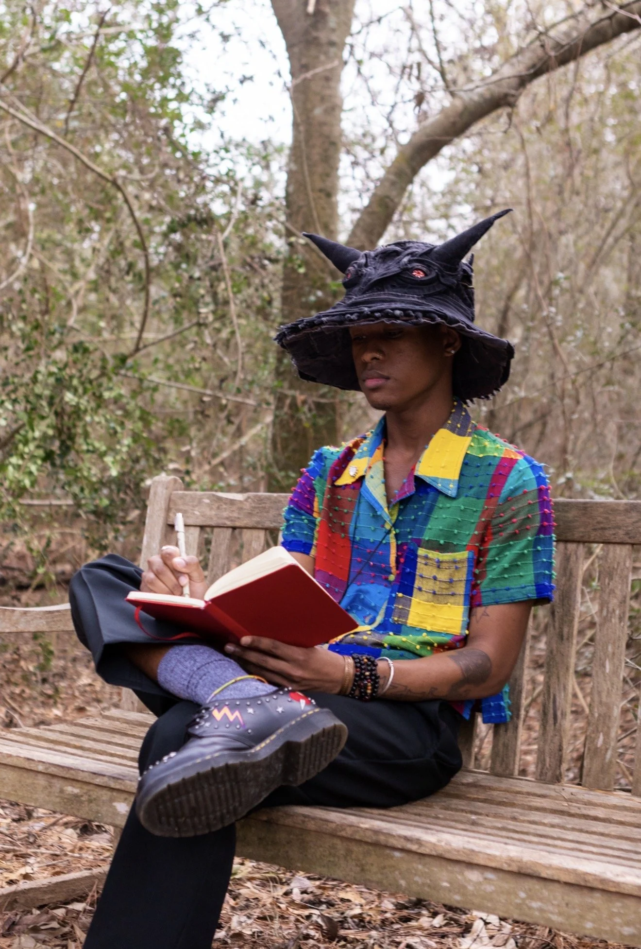 A young man sitting on a wooden park bench, reading a book with a red cover, wearing a colorful, patchwork shirt, a witch hat with horns, and black shoes, in a forest with trees and leaves on the ground.