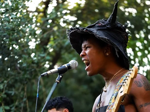 A woman wearing a witch hat singing into a microphone while playing a guitar outdoors.