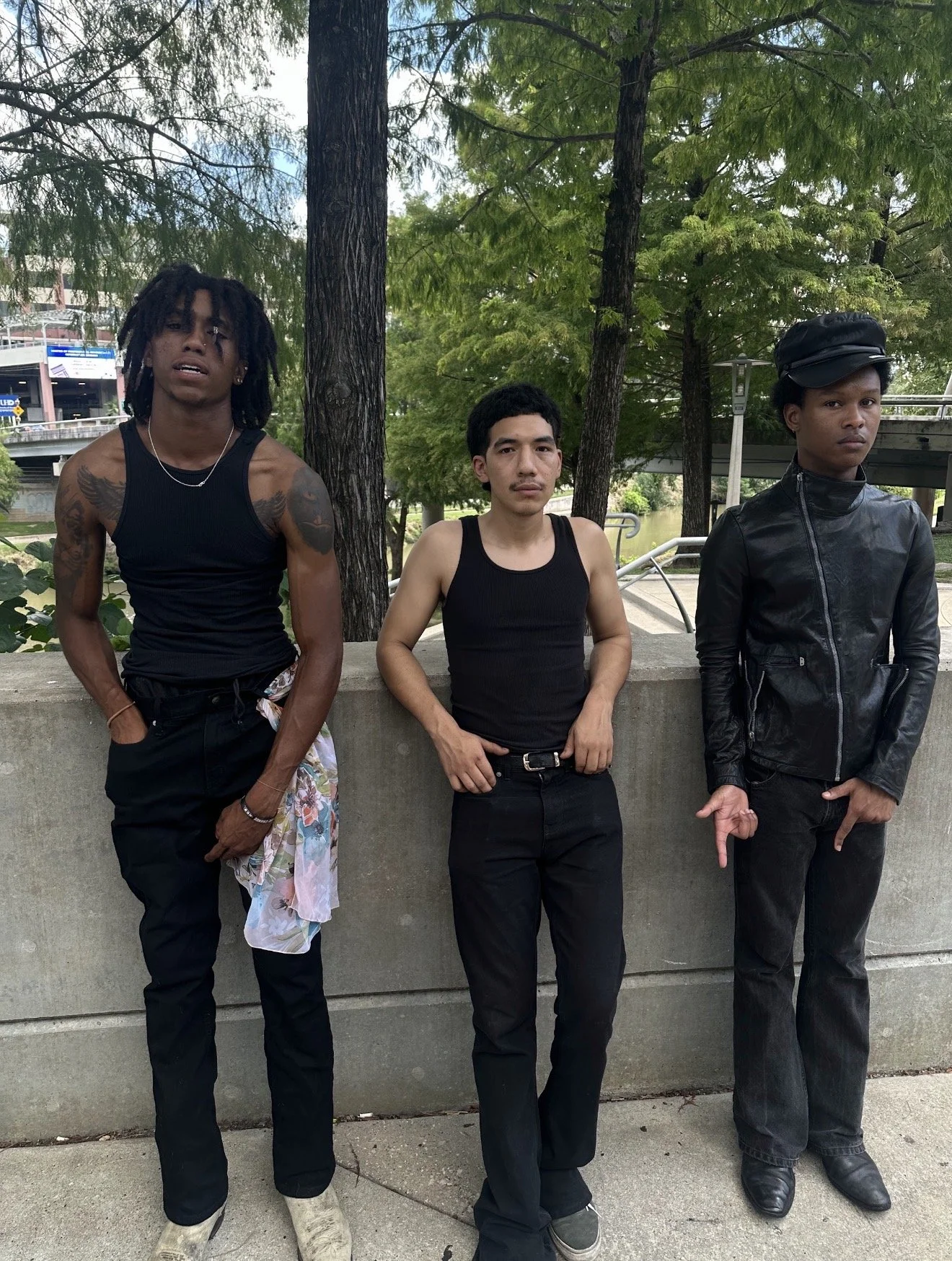 Three young men standing outdoors near a ledge with trees and a river behind them. The man on the left has dreadlocks, tattoos, and is wearing a sleeveless black shirt. The man in the middle has short black hair and is wearing a black tank top. The man on the right is wearing a black leather jacket, a black cap, and has short hair.