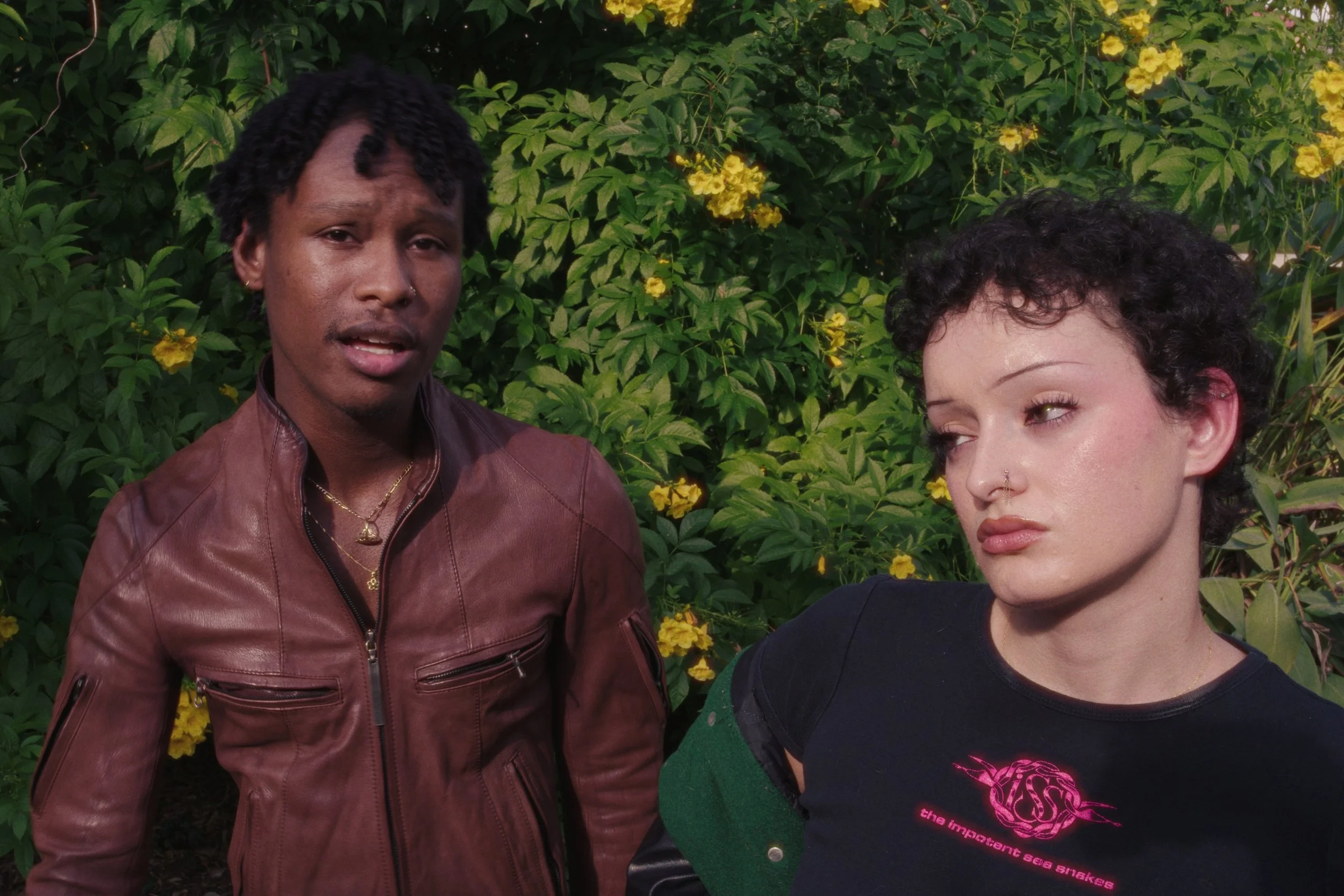 Two women standing outdoors in front of green foliage with yellow flowers. One woman has dark, curly hair and is wearing a black top, and the other has short, dark hair styled in twists and is wearing a brown leather jacket.