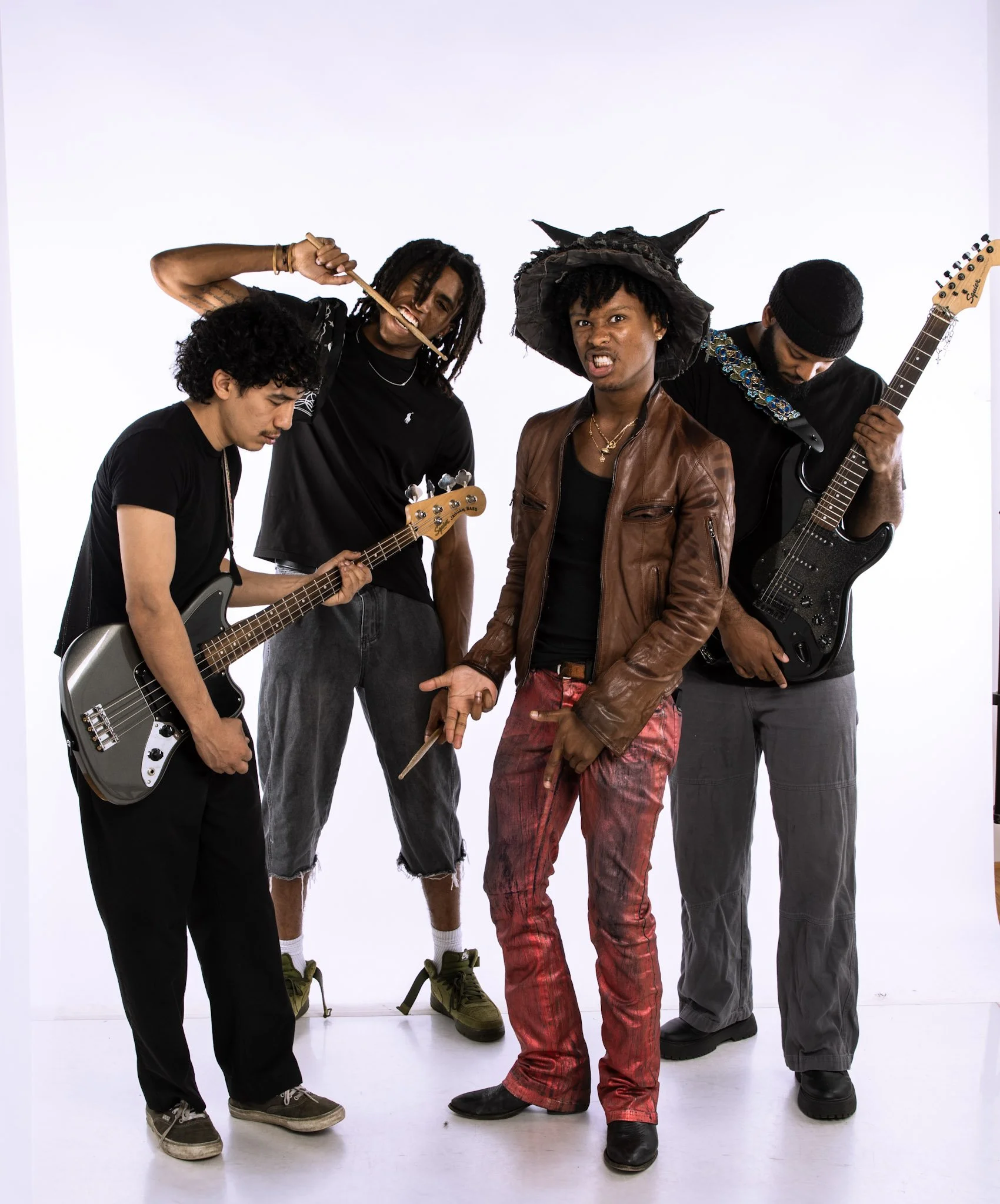 Five men are standing close together against a plain white background. They are dressed in casual and eclectic clothing, with some holding musical instruments like an electric guitar and a bass guitar. One man is wearing a large, pointed hat with horns, and another is wearing a knit beanie. They display expressive facial expressions and poses conveying attitude and energy.