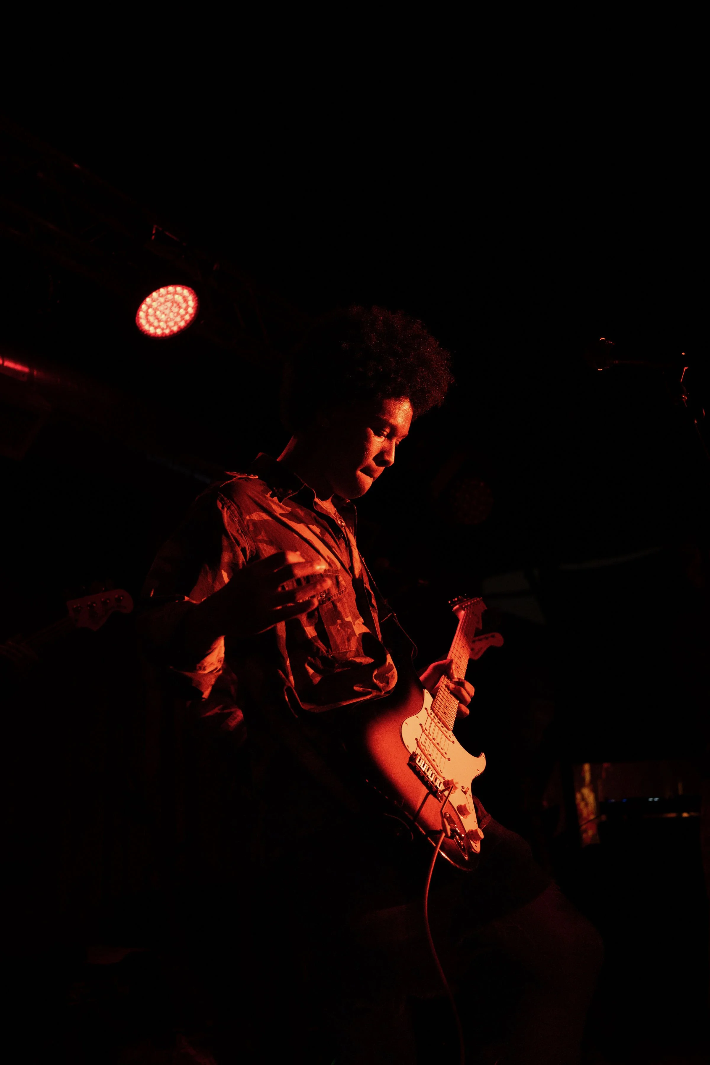 A musician playing an electric guitar on stage, illuminated by red stage lights against a dark background.