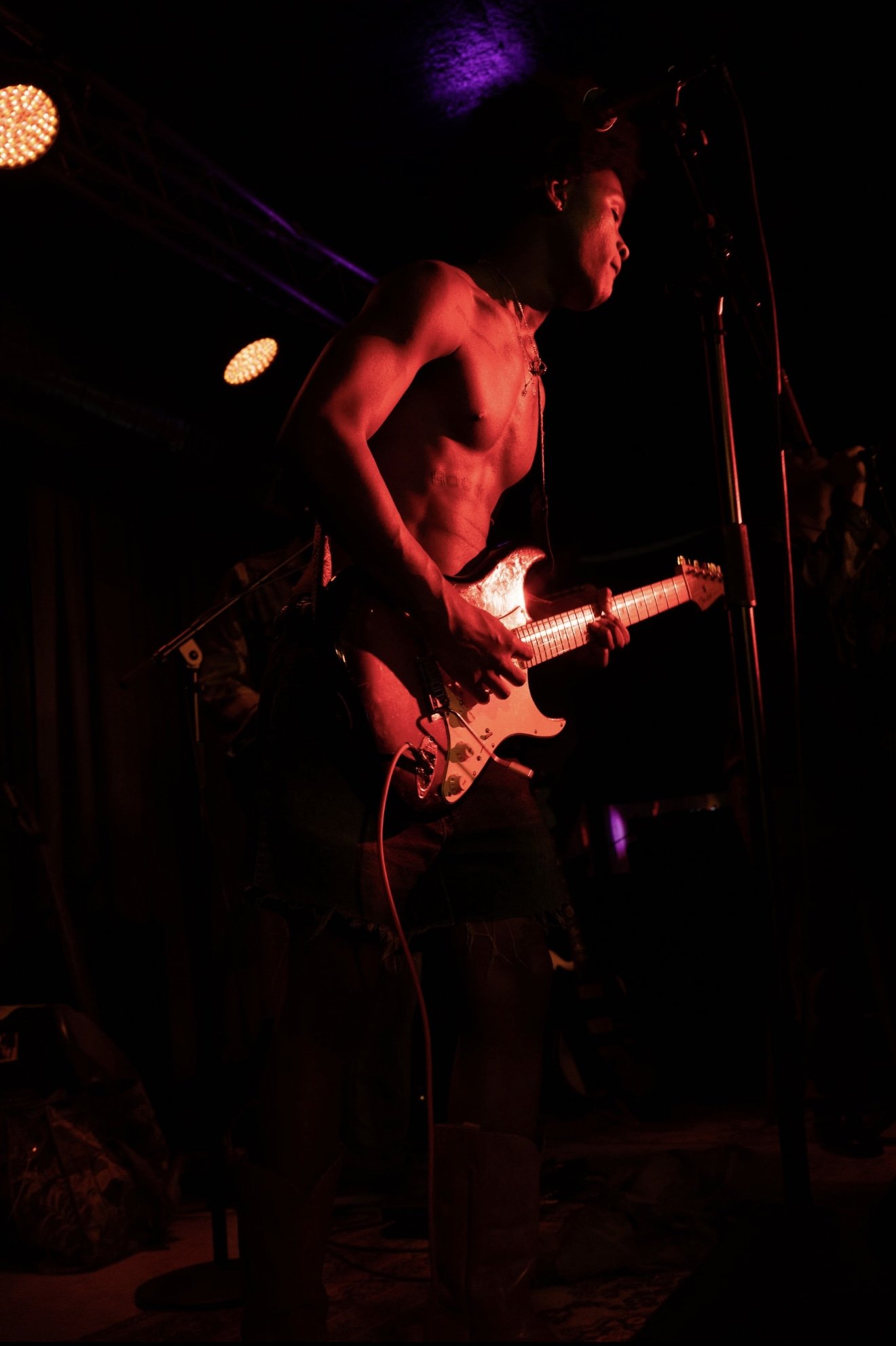 A shirtless male musician playing an electric guitar on stage during a performance, with red and purple stage lighting and dark background.