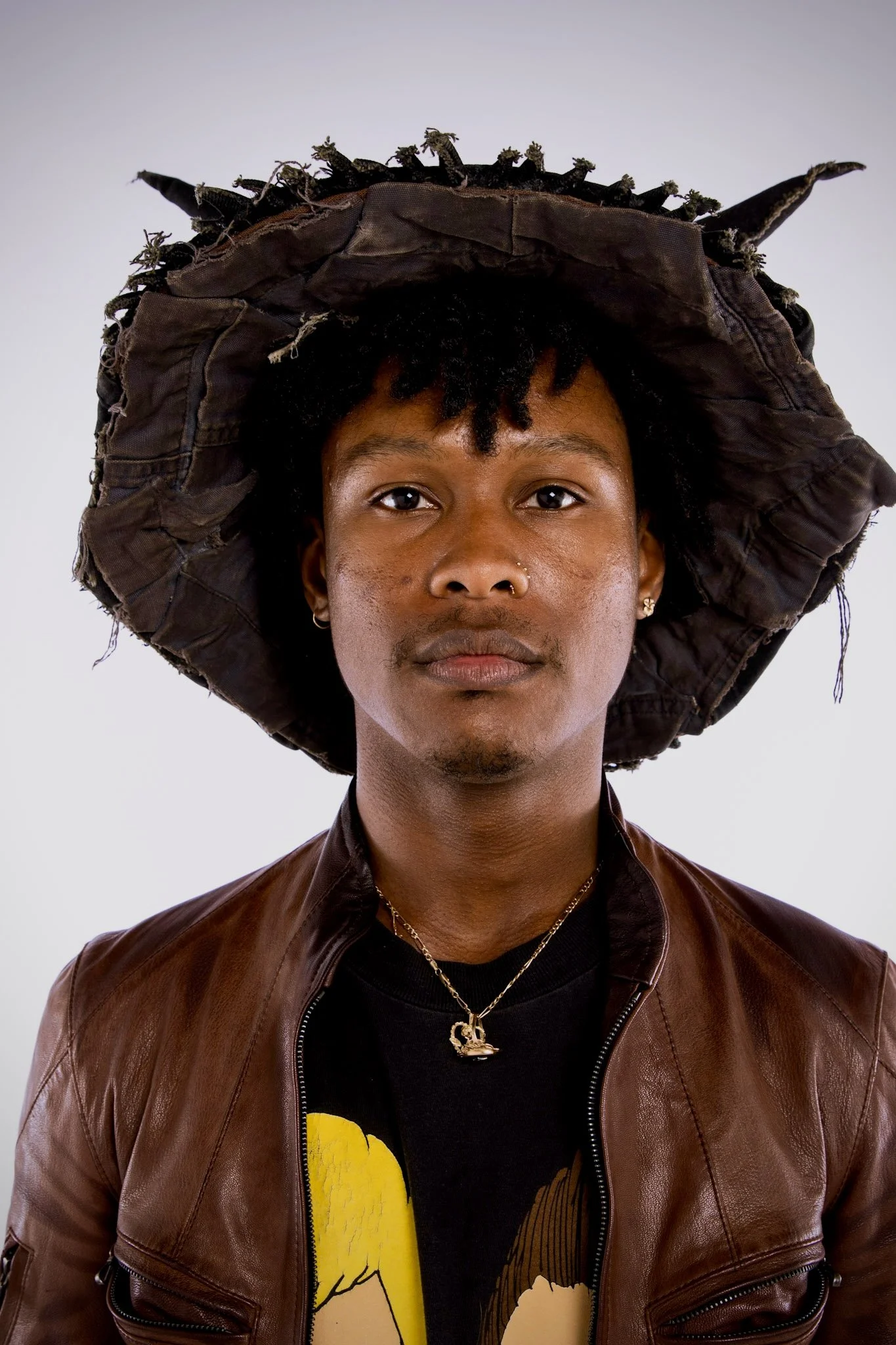A person with short, curly black hair wearing a large, worn, dark hat with twisted points, a brown leather jacket, and layered jewelry, including a gold necklace with a necklace pendant.