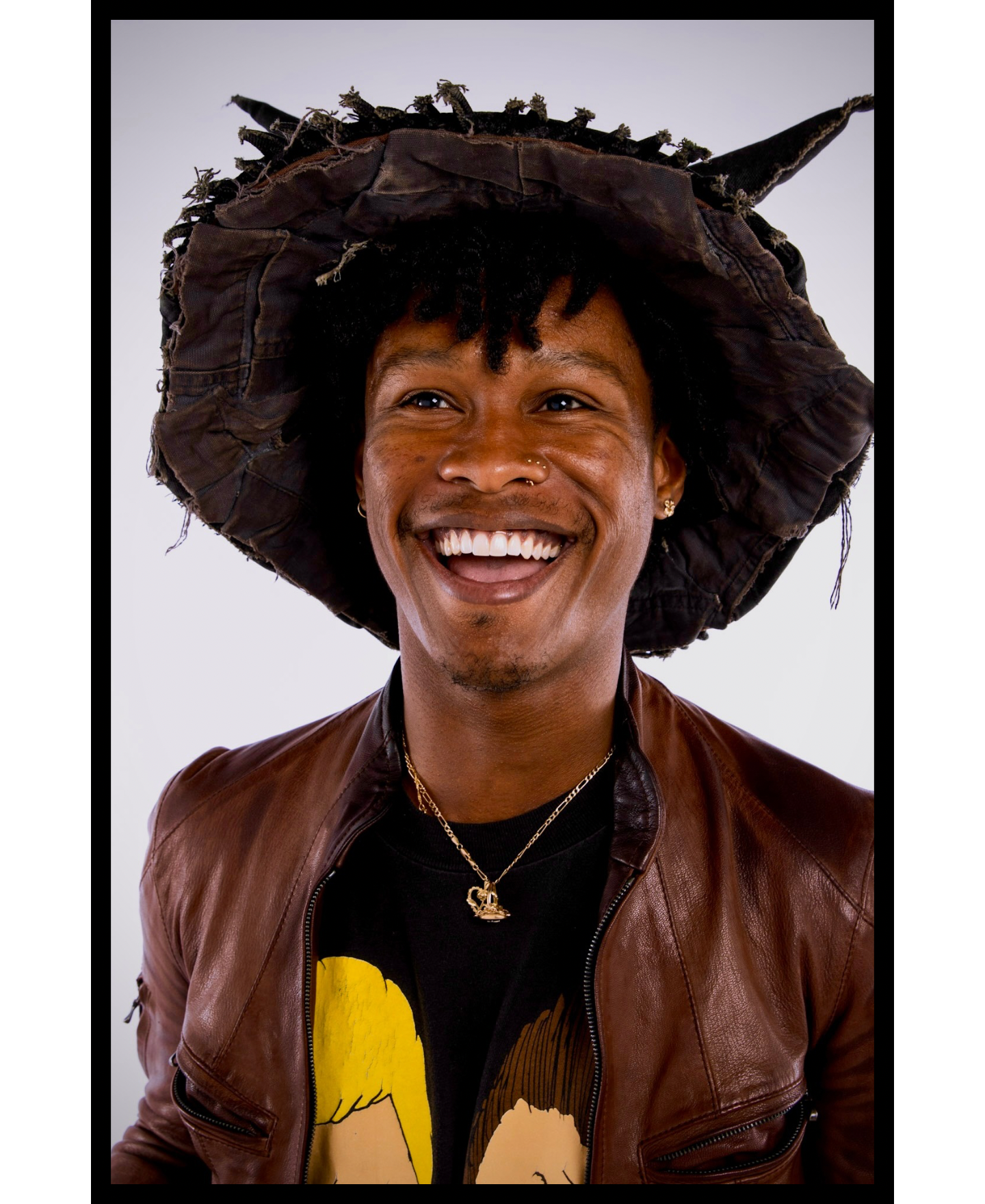 A smiling person with short curly hair wearing a large weathered hat with horns, a brown leather jacket, a black t-shirt with a cartoon character, and gold jewelry, posing against a plain white background.