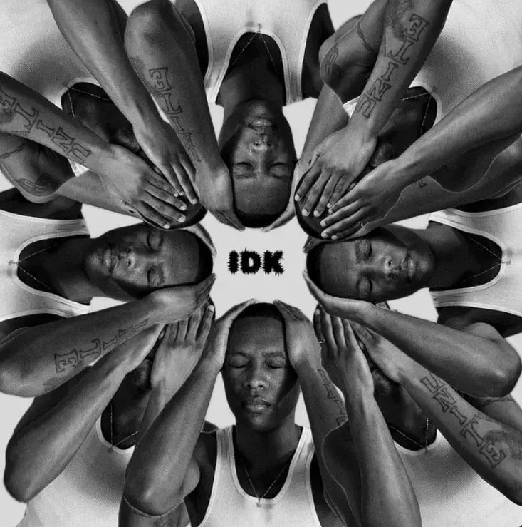 A black and white photo of eight women with hands over their ears, forming a circle. They have closed eyes and tattoos on their arms, and are wearing sleeveless shirts. In the center, the word "IDK" is written.