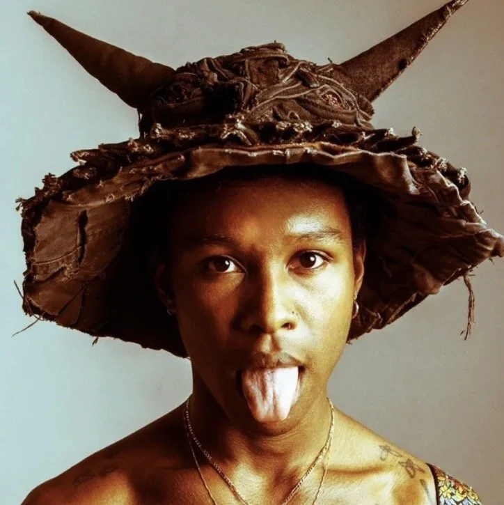 A person with dark skin wearing a large, pointed, brown hat resembling a witch's hat made of rough, textured material, and sticking out their tongue.