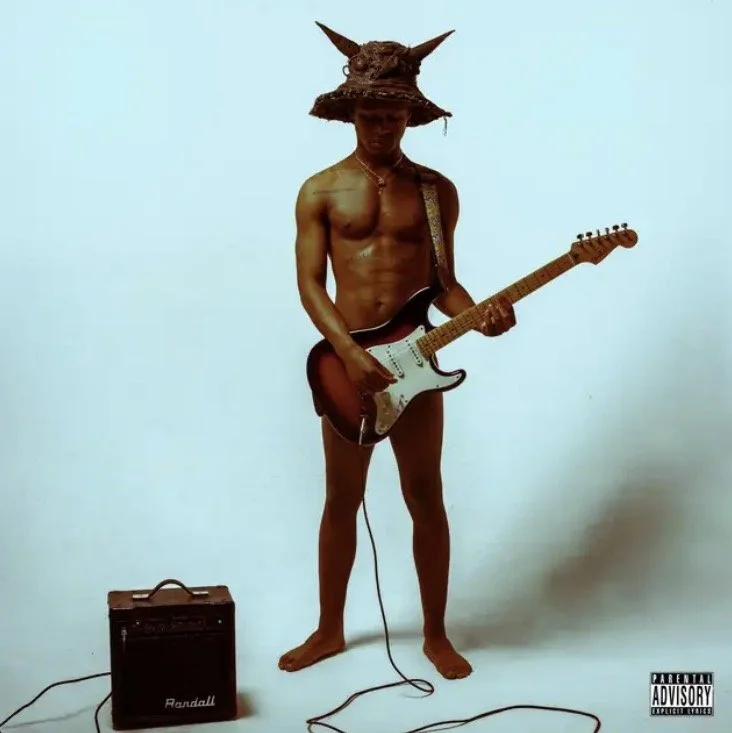 A shirtless man wearing a wide-brimmed hat with pointed ears, playing an electric guitar outdoors on a plain background with an amplifier on the ground.