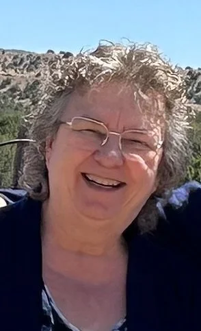 Portrait of artist Sharon Marrell smiling outdoors with trees and a rocky hillside in the background.