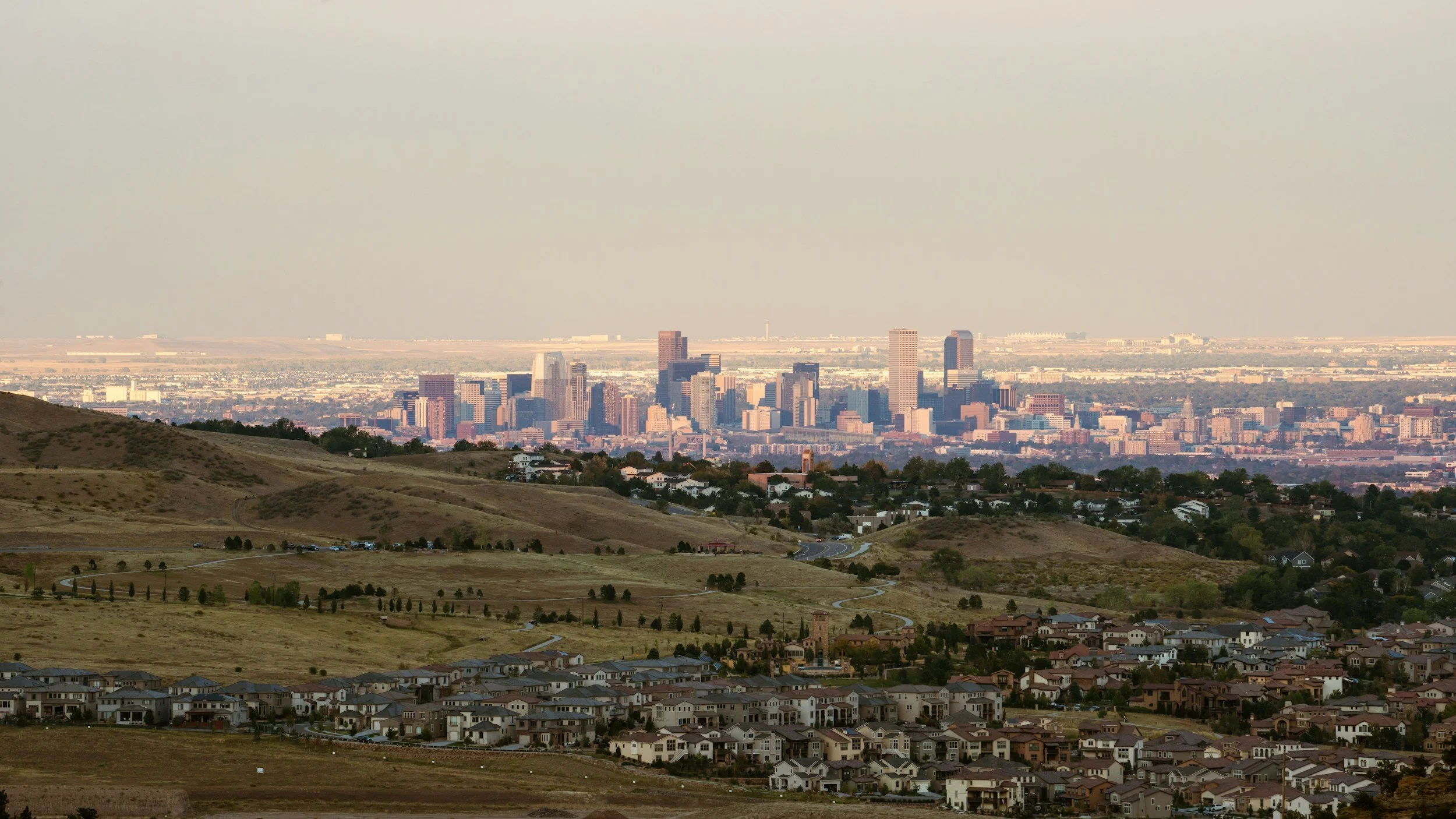 City skyline with tall buildings in the distance, rolling hills and residential houses in the foreground.