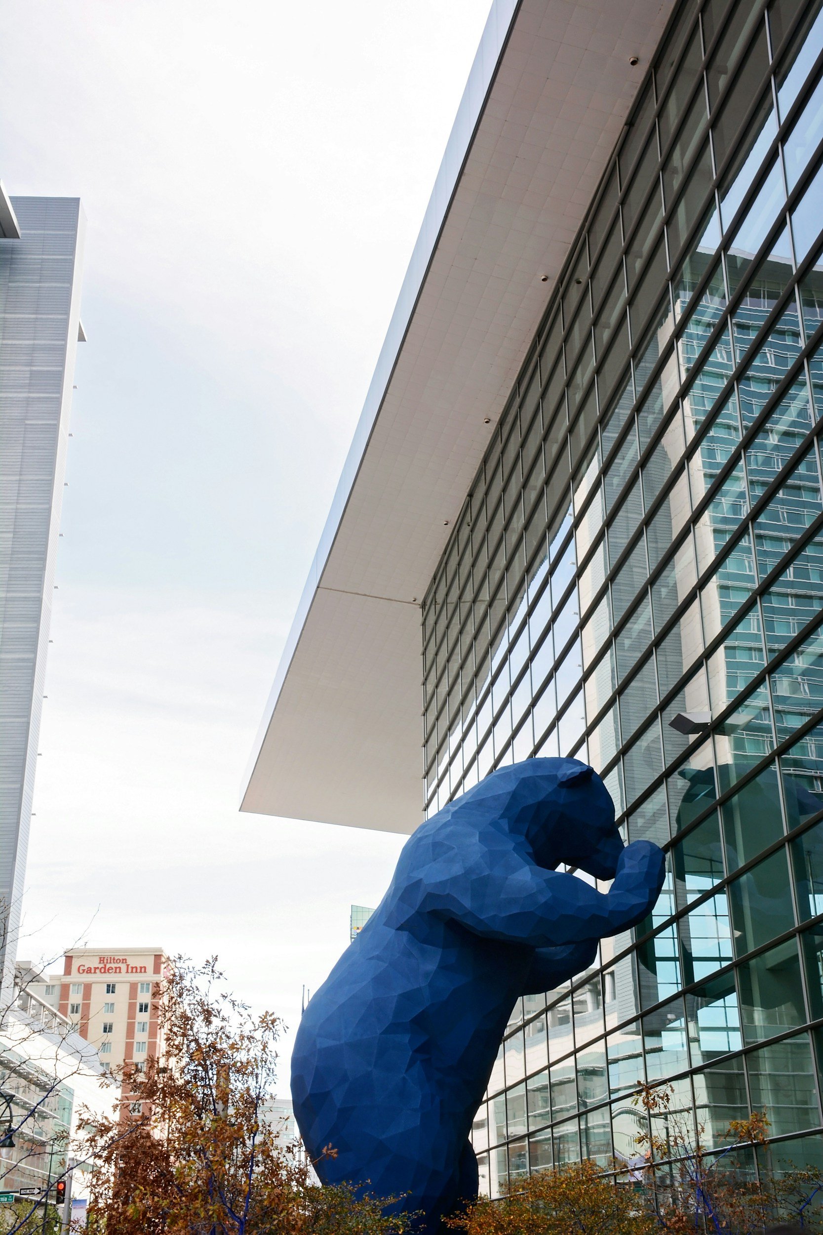 A large blue sculpture of a person leaning against a modern glass building with a white overhang, with trees and a Hilton Garden Inn hotel in the background.