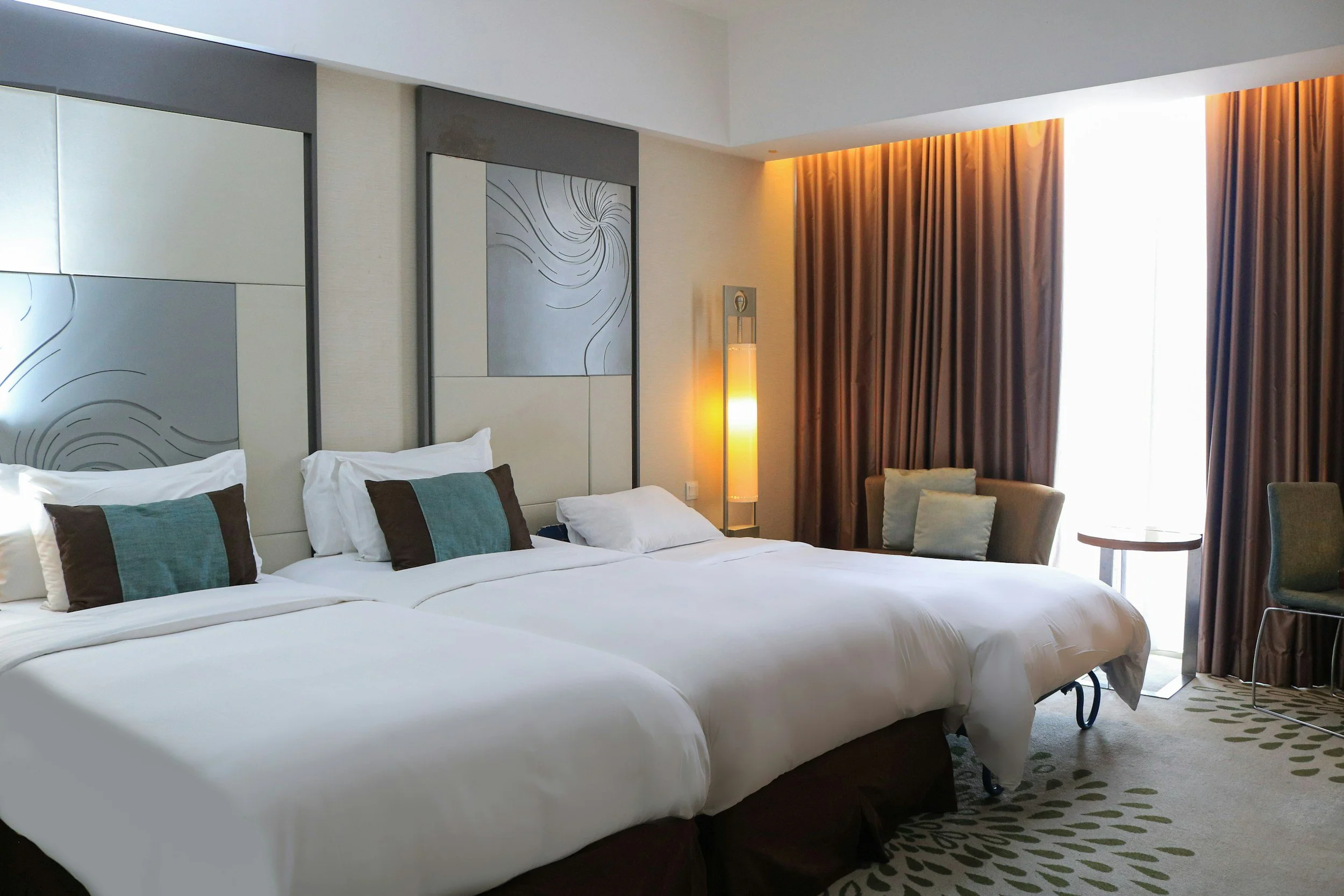 A hotel room with two large beds, decorative pillows, a sitting area with chairs, window with brown curtains, and a lampshade near the window.
