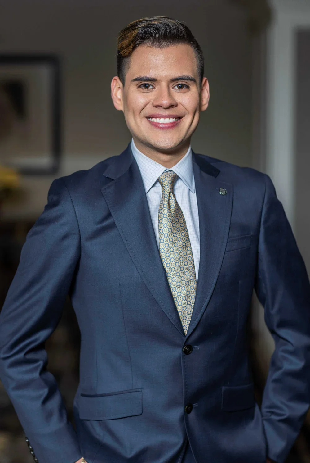 A young man with short, dark hair, wearing a navy blazer, a light blue dress shirt, and a patterned yellow tie, smiling confidently in a professional setting.