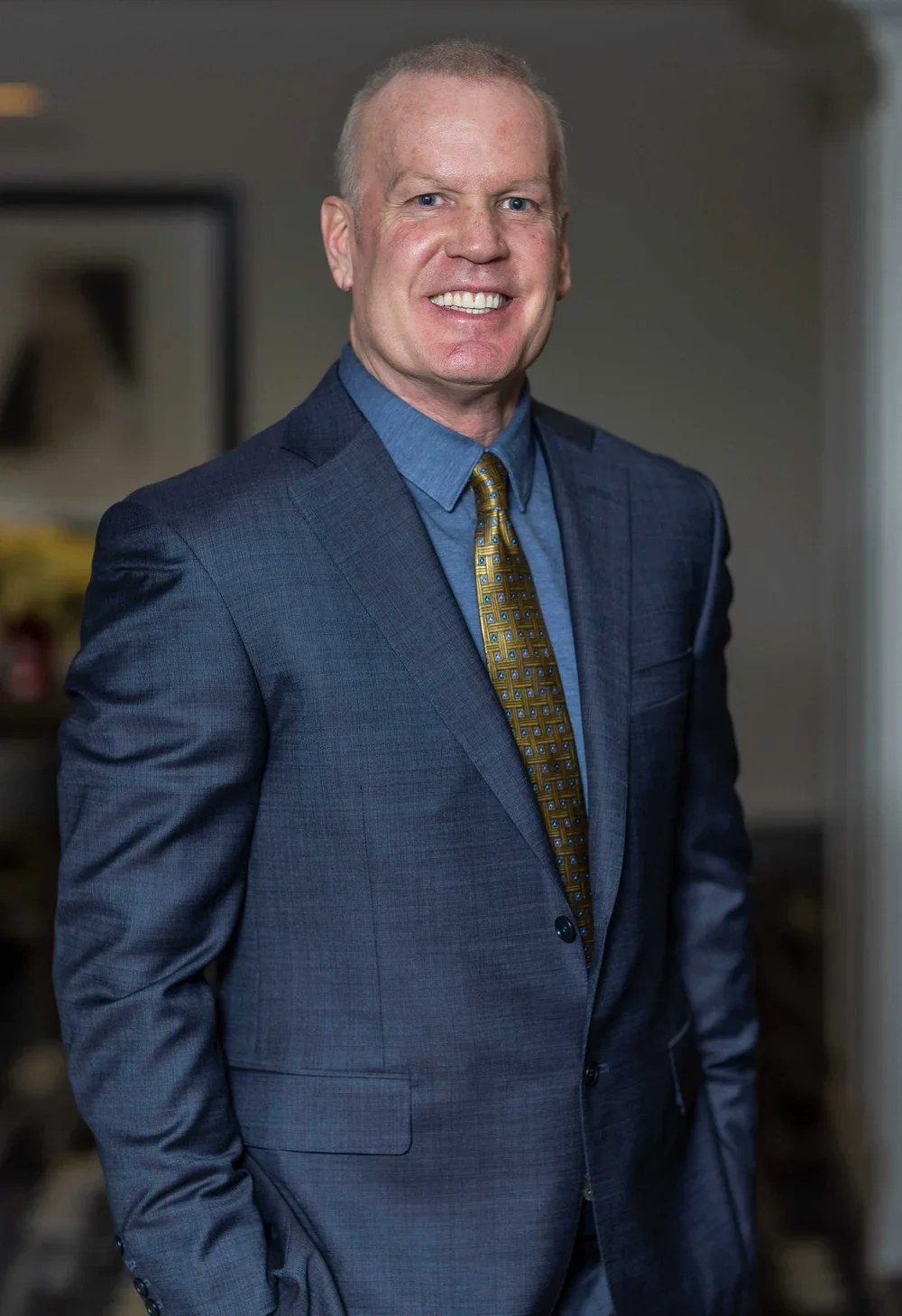 A professional man in a dark blue suit, blue shirt, and patterned tie, smiling and standing indoors.