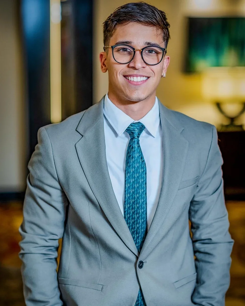 A young man with short dark hair, glasses, and earrings, wearing a light gray suit jacket, a white shirt, and a blue patterned tie, smiling in an indoor setting.