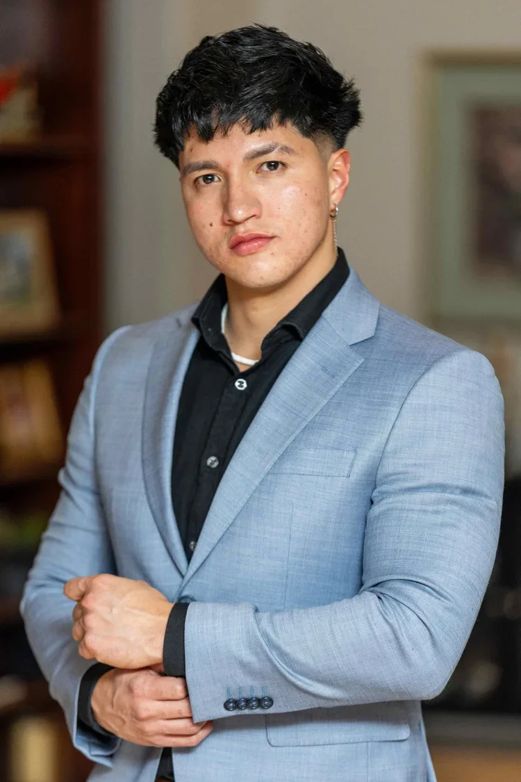 Portrait of a young man wearing a light gray blazer and black shirt, with short black hair, in an indoor setting with blurred background, possibly in a home or office.
