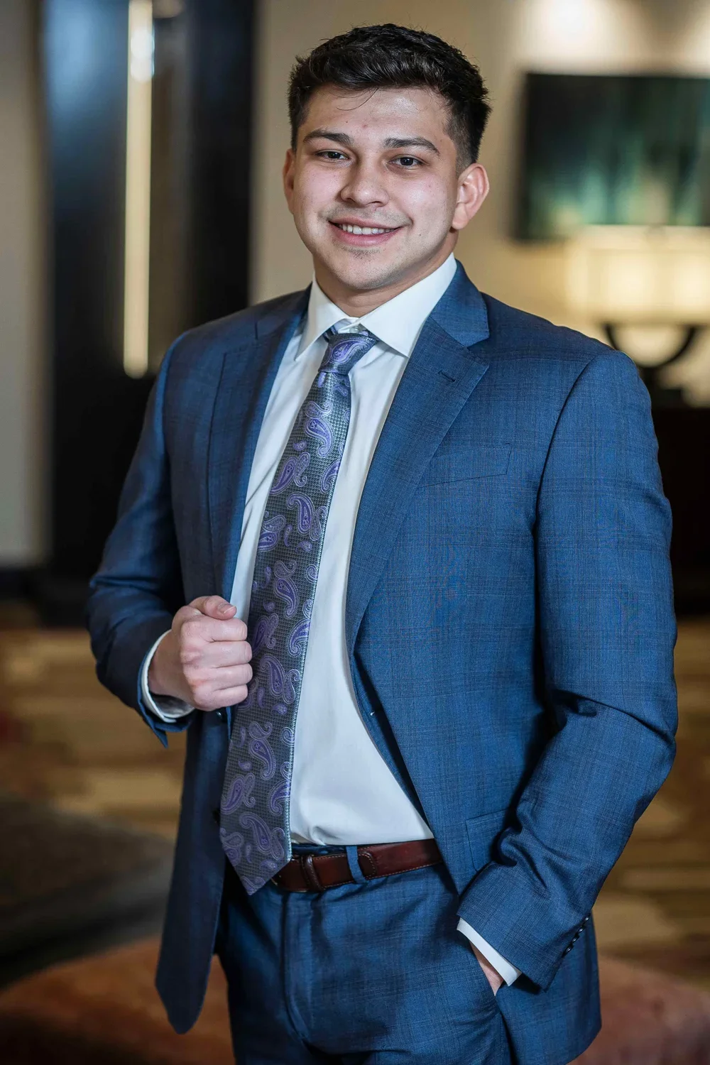 A young man in a blue suit, white shirt, and patterned tie standing indoors, smiling with one hand adjusting his lapel.