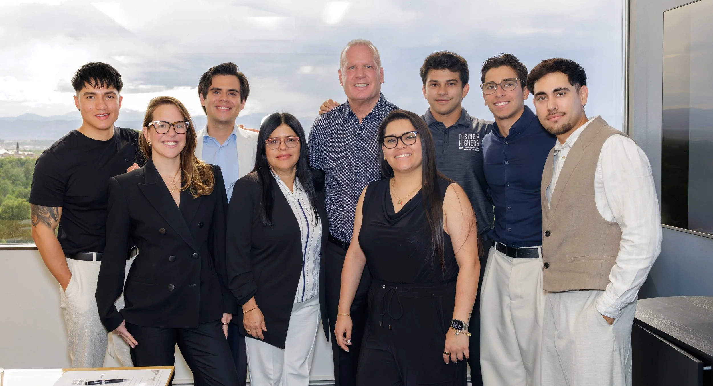 A diverse group of ten people, including men and women, standing together indoors with a window overlooking a cityscape and mountains in the background, smiling at the camera.