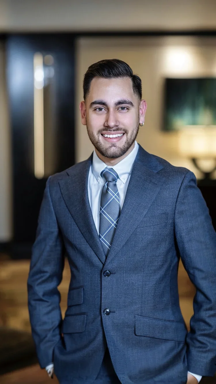 A young man with dark hair and a beard wearing a blue suit, white shirt, and a patterned tie, smiling and standing in an indoor setting.