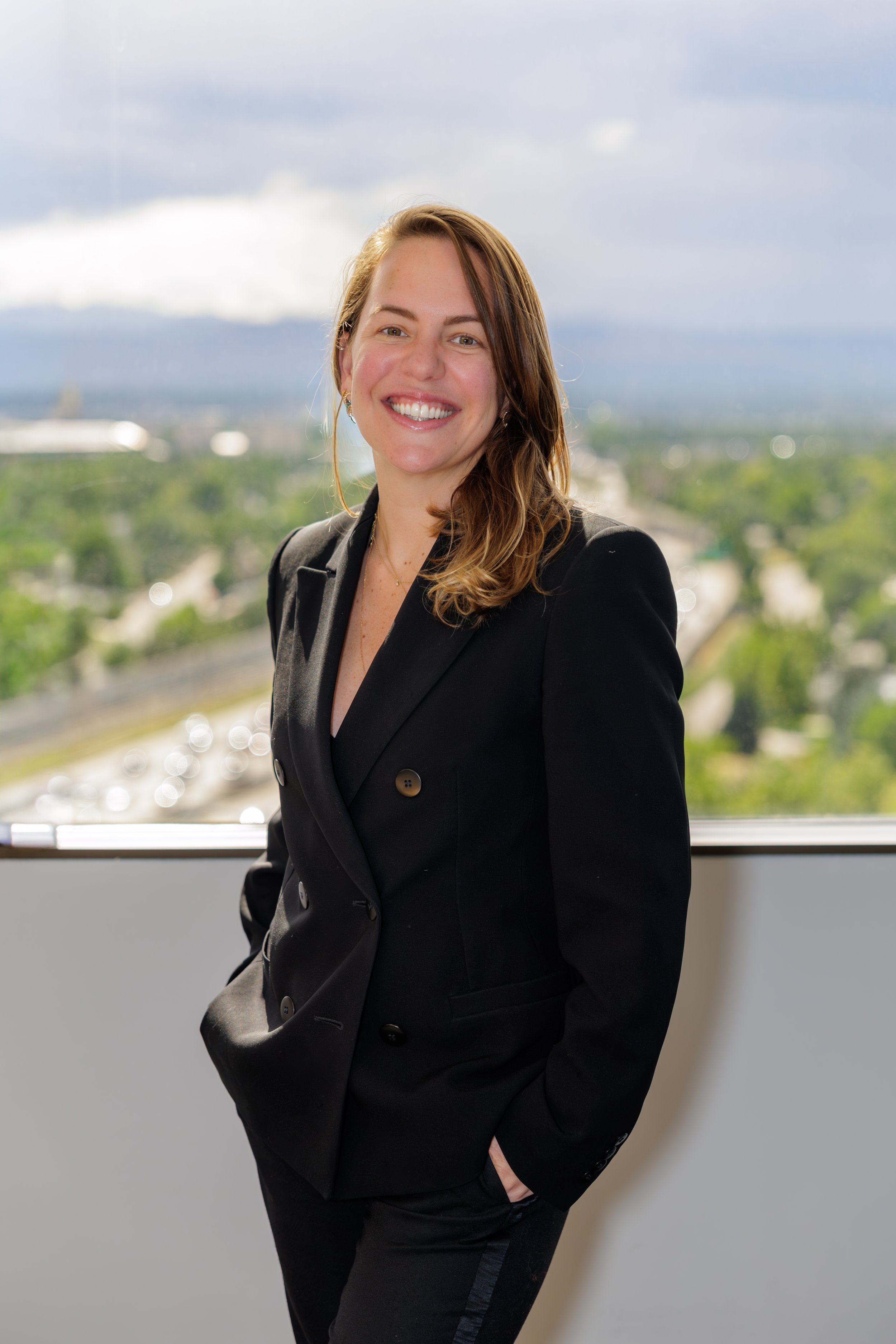 A smiling woman with shoulder-length blond hair, dressed in a black blazer and black pants, standing indoors near a window with a cityscape and cloudy sky in the background.