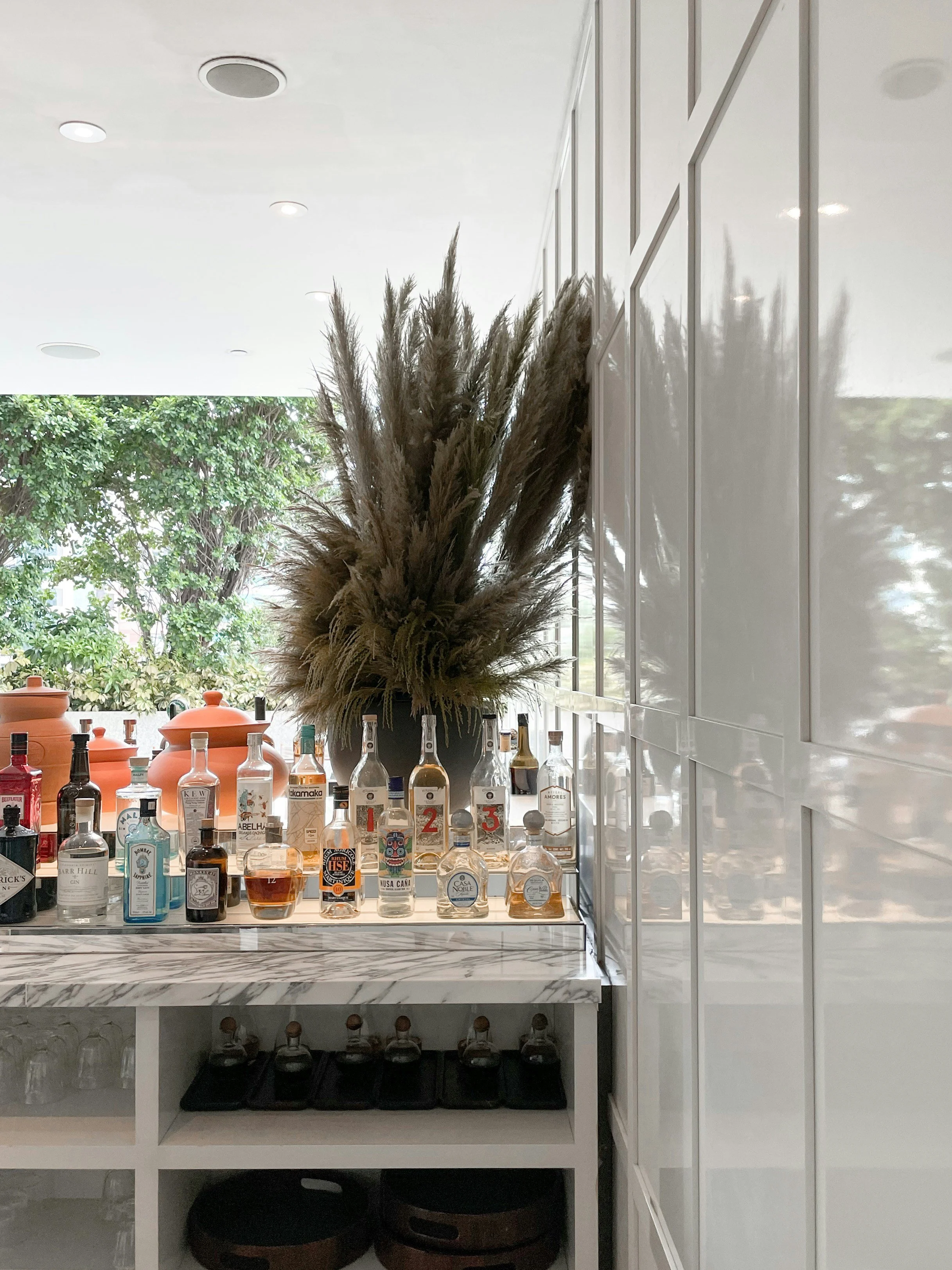 A bar counter with various liquor bottles, a large decorative dried plant in a vase, and storage containers underneath, with a bright window and white cabinetry in the background.