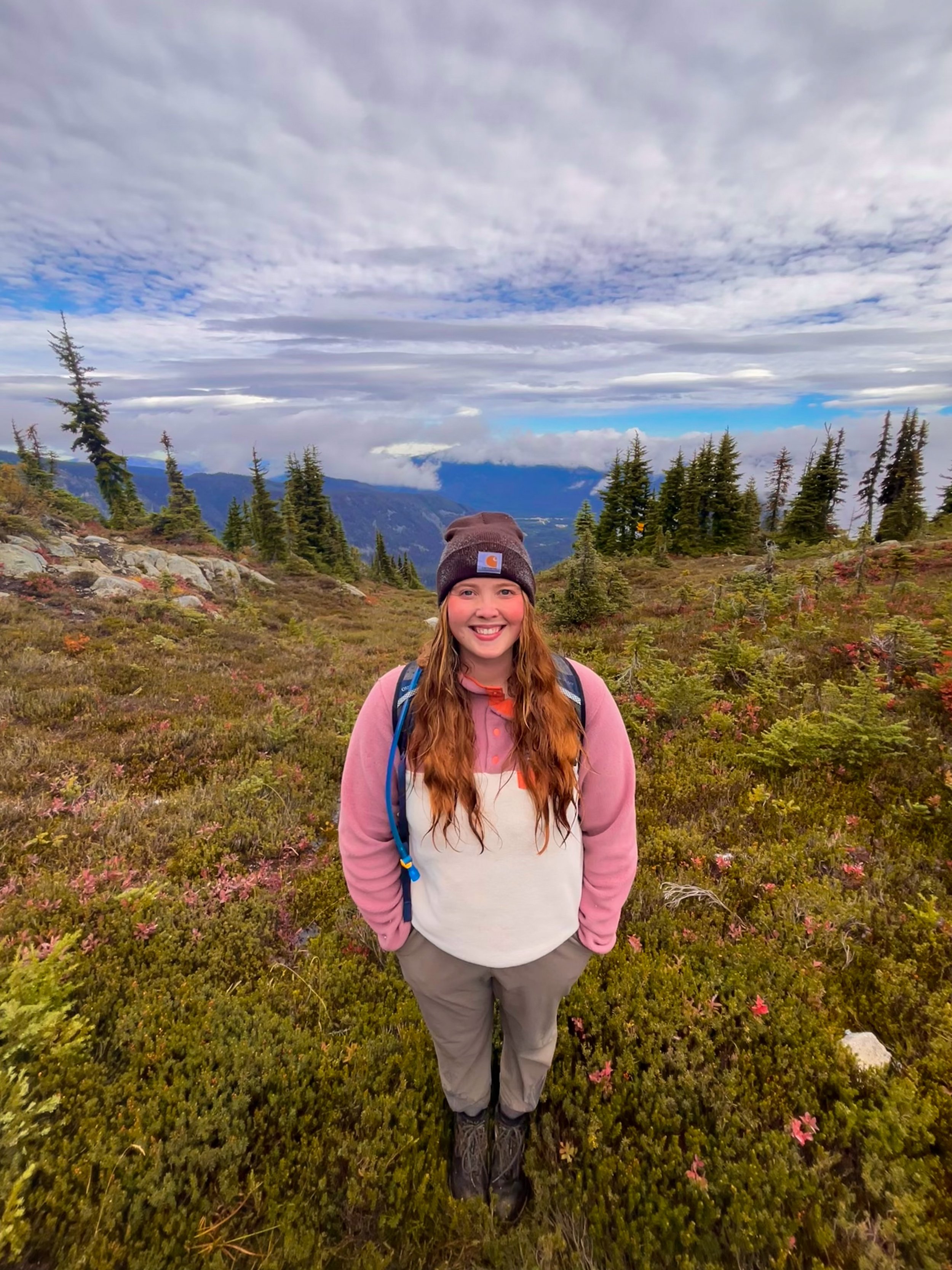 A smiling woman wearing a beanie, pink jacket, and hiking gear standing in a lush mountain landscape with trees and cloudy sky.