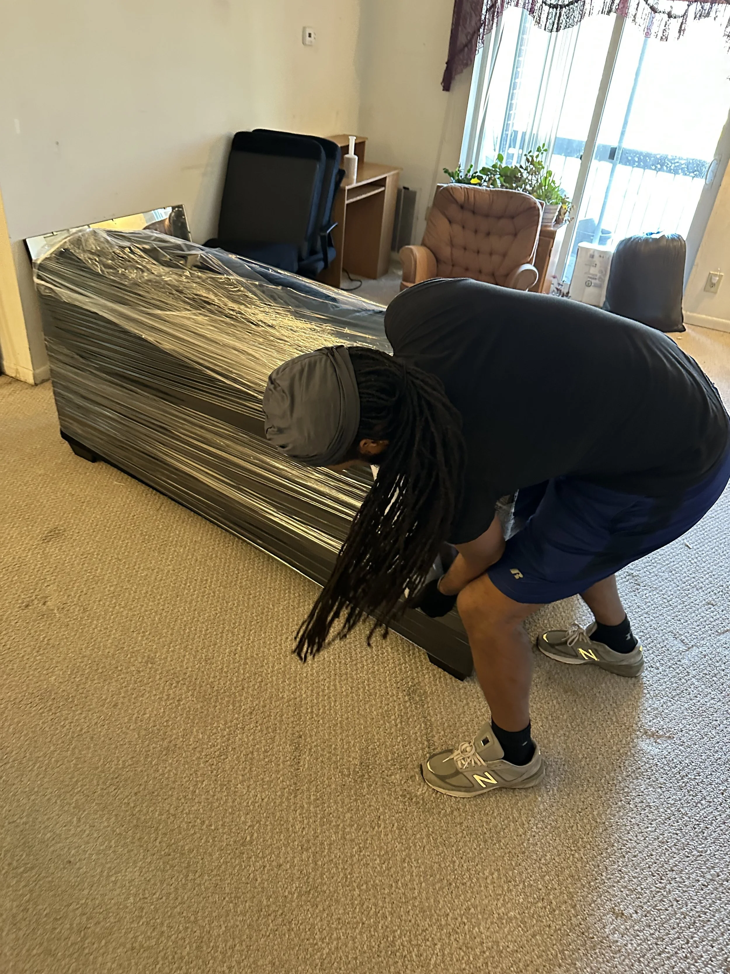 A person with long dreadlocks is bending over a couch wrapped in plastic, wearing sneakers, shorts, and a T-shirt.