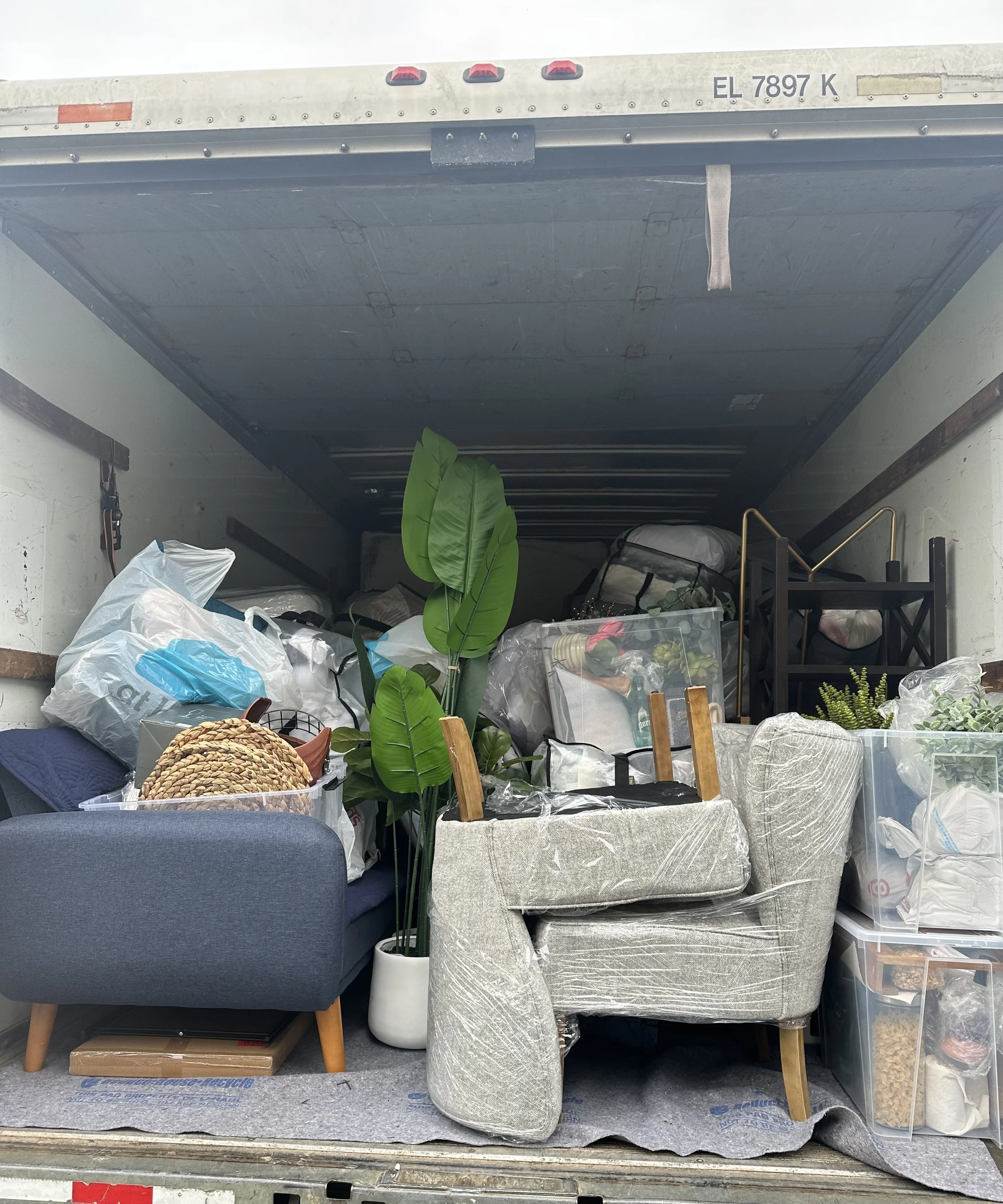 The image shows a storage unit or truck bed filled with various items, including large wrapped packages, plastic containers, furniture pieces, plants, and miscellaneous household items, with a mostly white interior and some visible objects on or under the items.