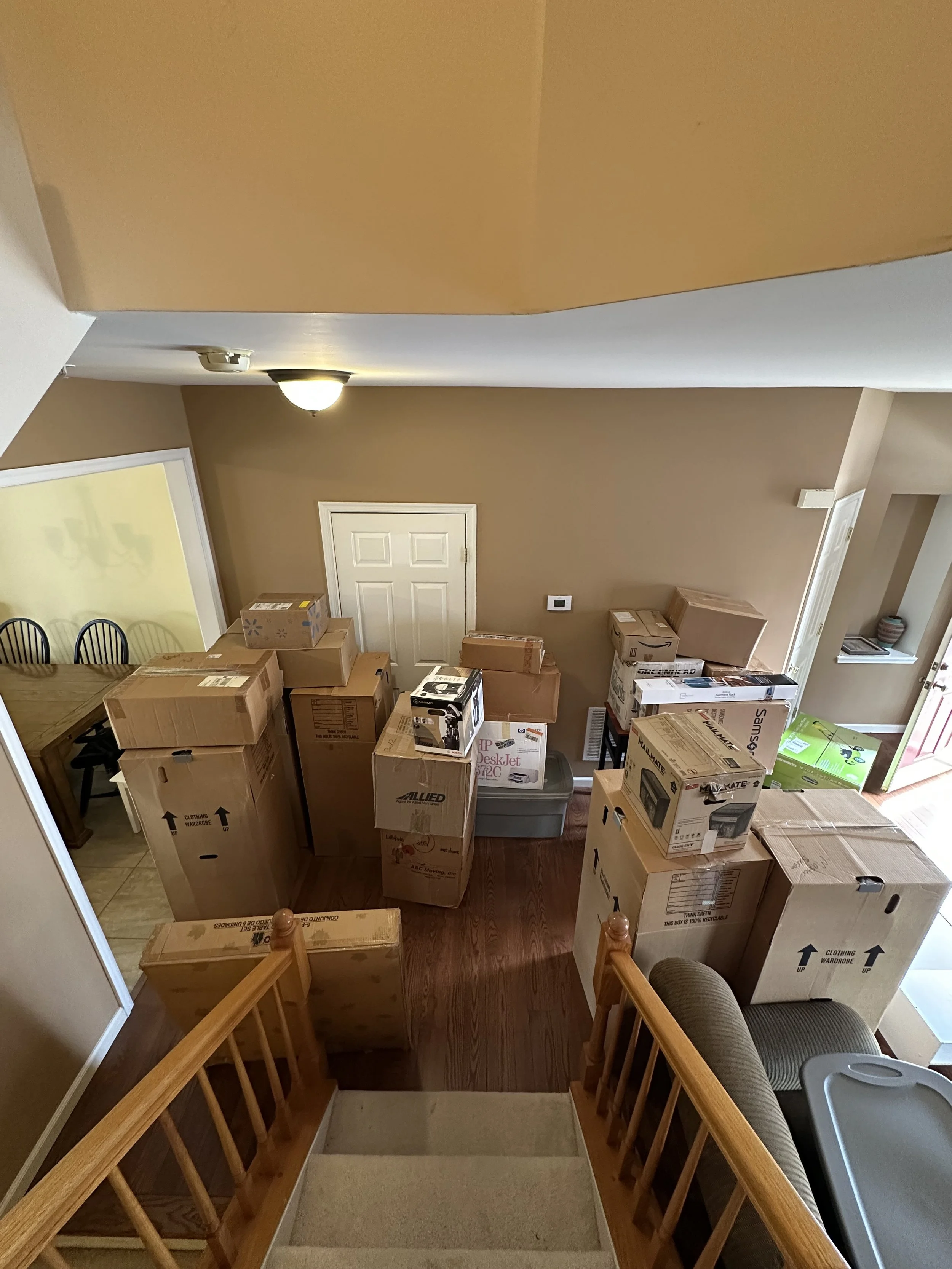 A staircase viewed from the top, leading down into a room filled with packed moving boxes, some on the floor and some on a table, with a nearby dining area with chairs and a wooden table, beige walls, a door at the back, and a ceiling light.
