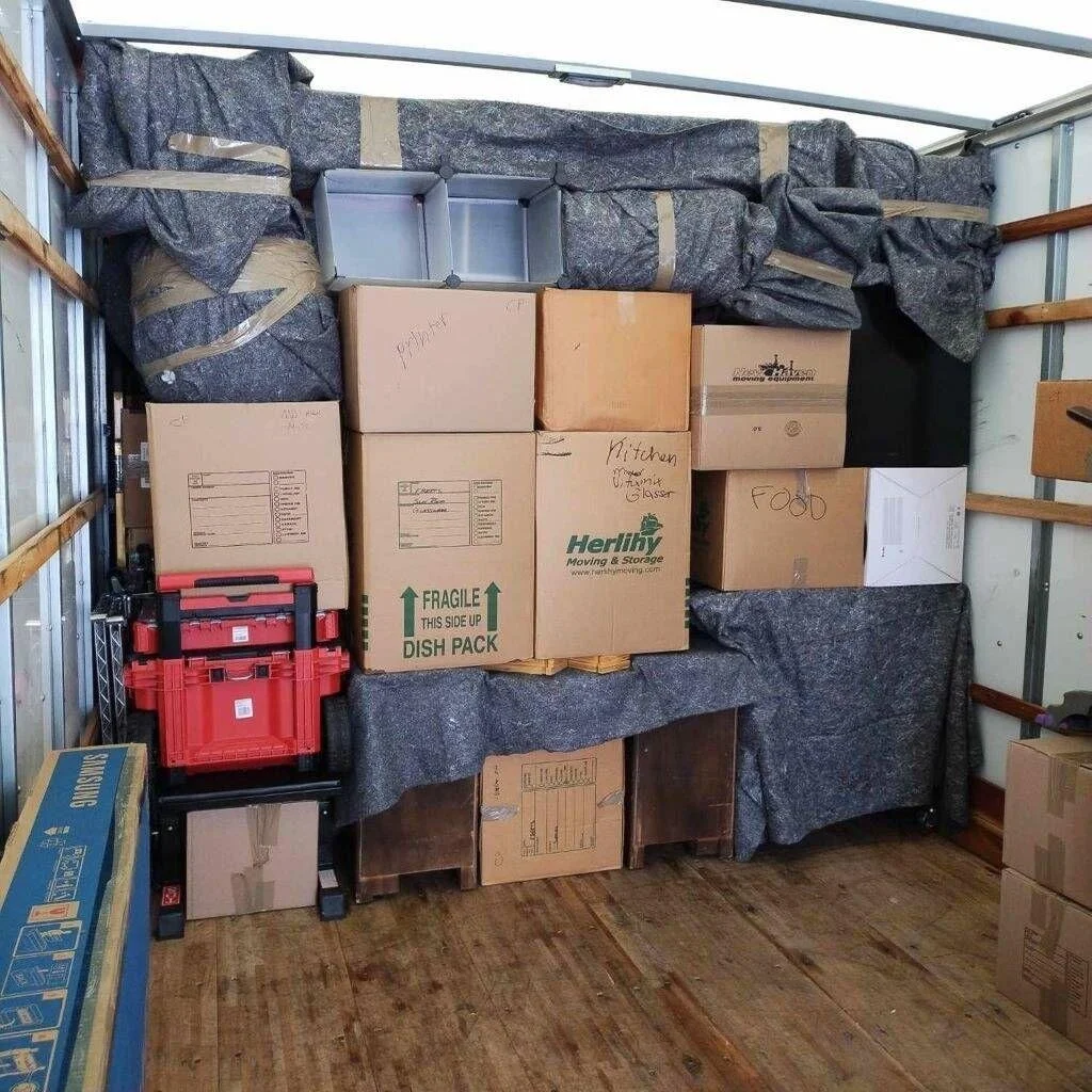 Packed moving truck with boxes labeled kitchen, food, and miscellaneous, some wrapped in gray packing material, along with red and blue storage containers and some wooden furniture.