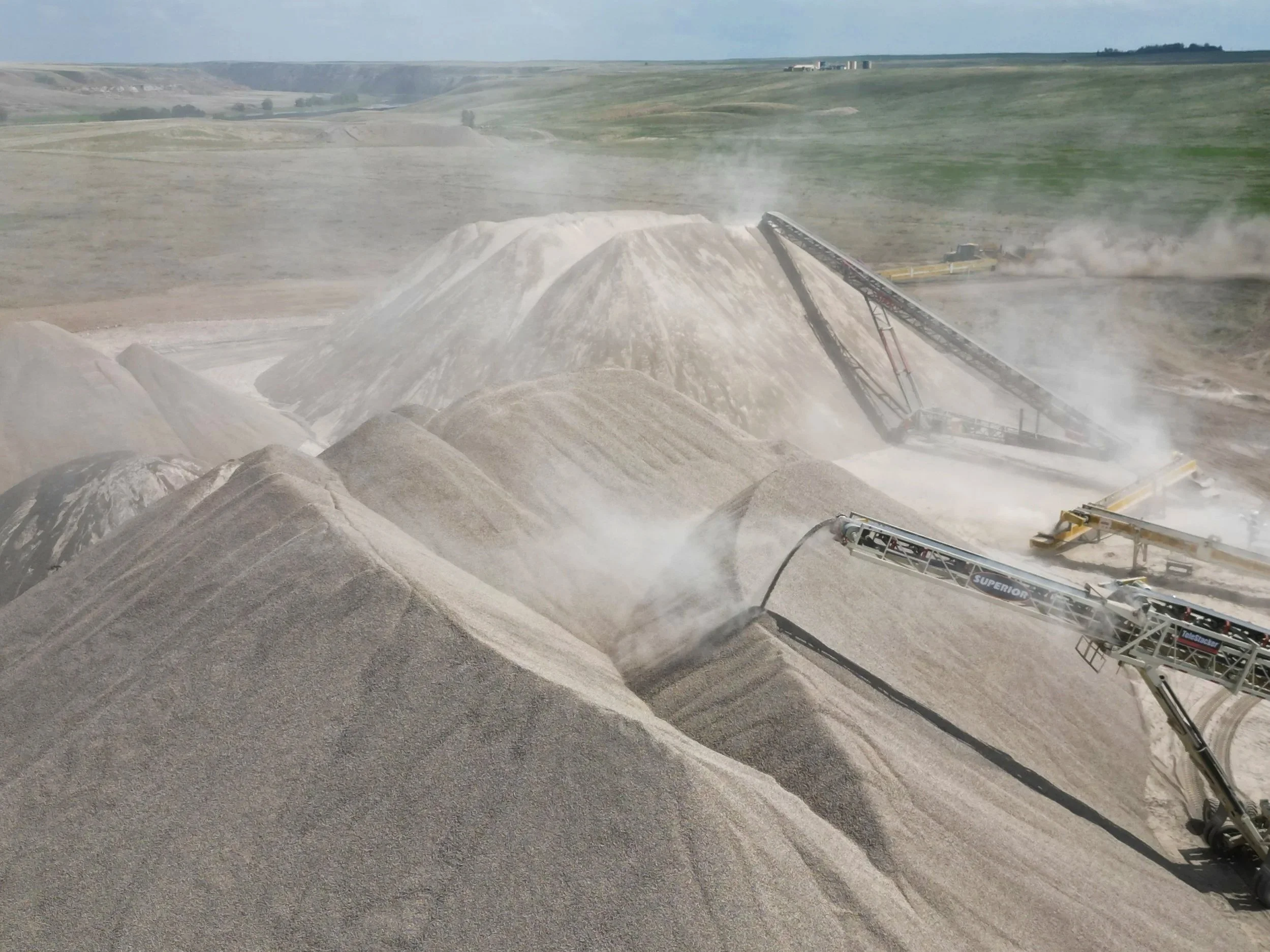A large quarry operation with a conveyor system moving gravel or similar material. The landscape is open with rolling hills and few trees in the background, under a clear sky.