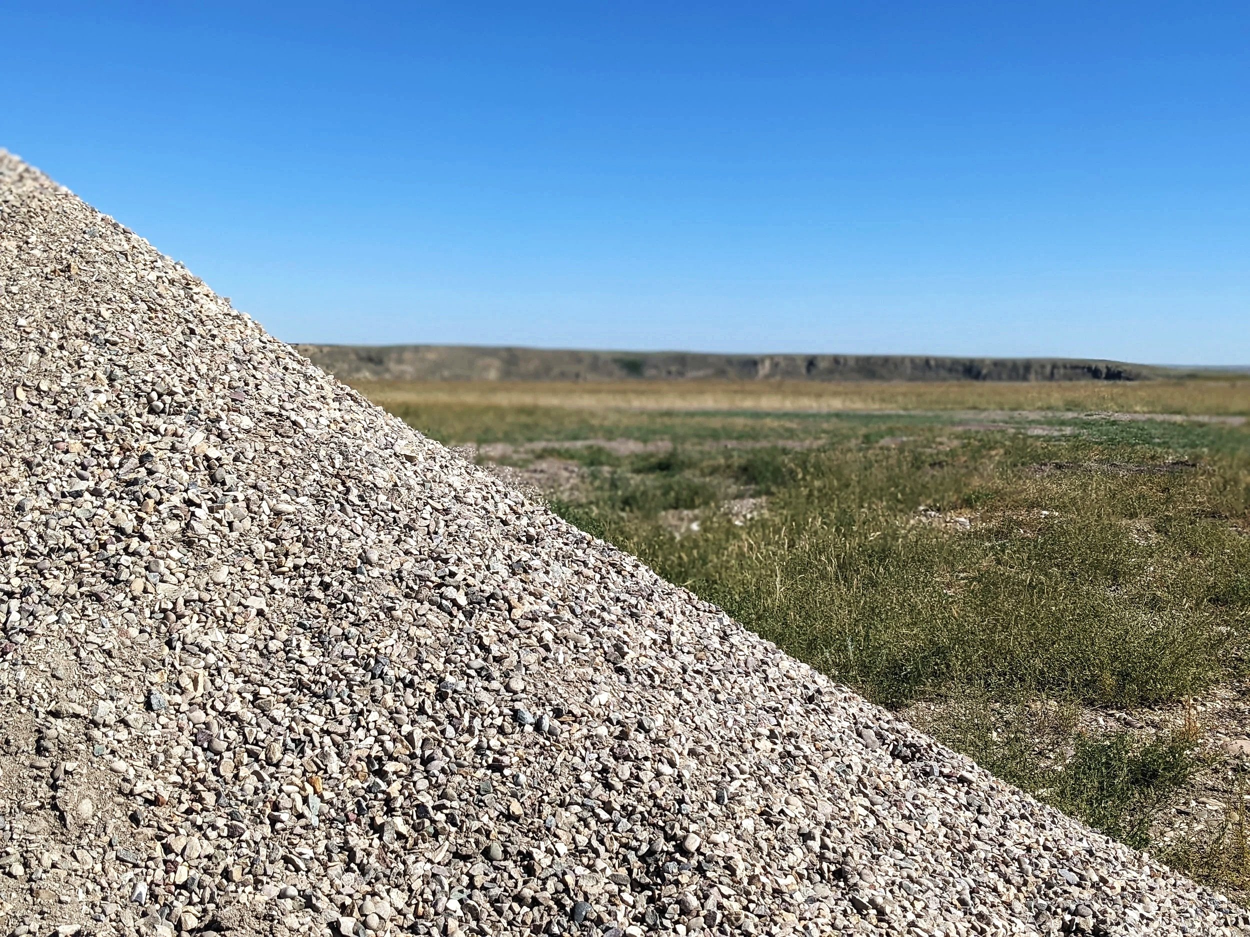 A large pile of gravel or small rocks in the foreground with a flat, grassy landscape and clear blue sky in the background.
