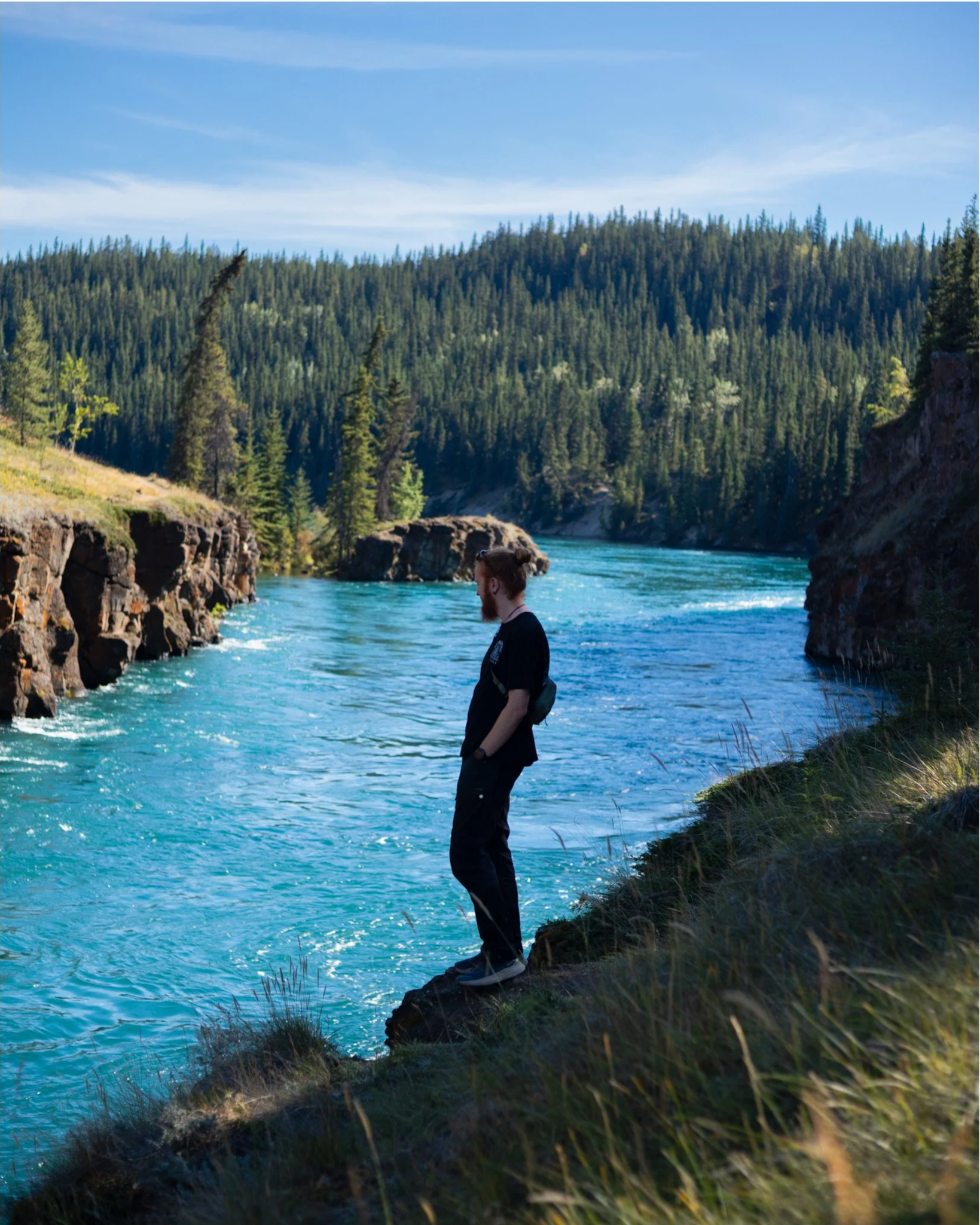 A man with a bun hairstyle and a beard, wearing a black shirt, standing on a rocky ledge beside a river surrounded by dense green forest and hills under a blue sky with wispy clouds.