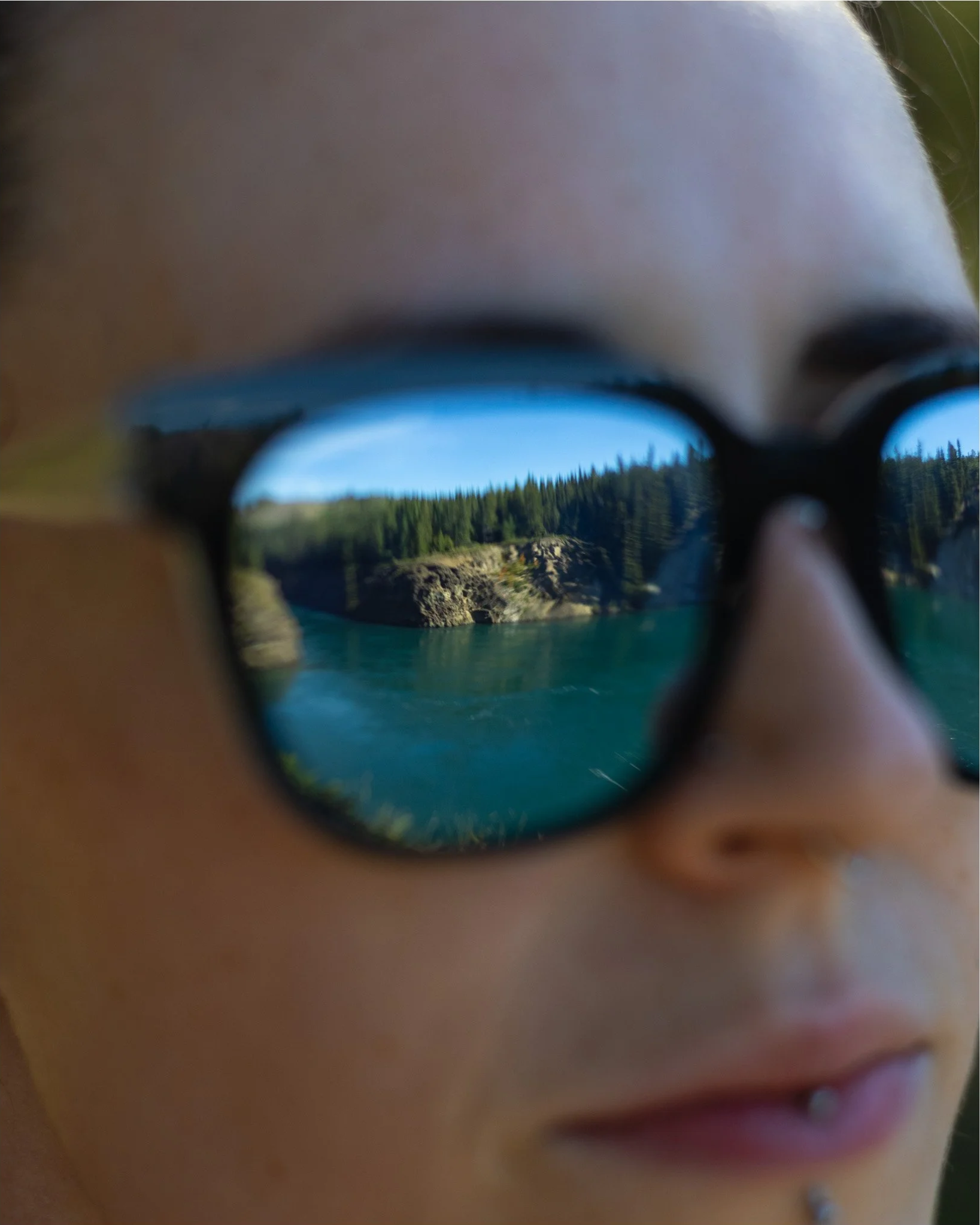 Close-up of a person wearing black glasses, reflecting a landscape with trees and a body of water.