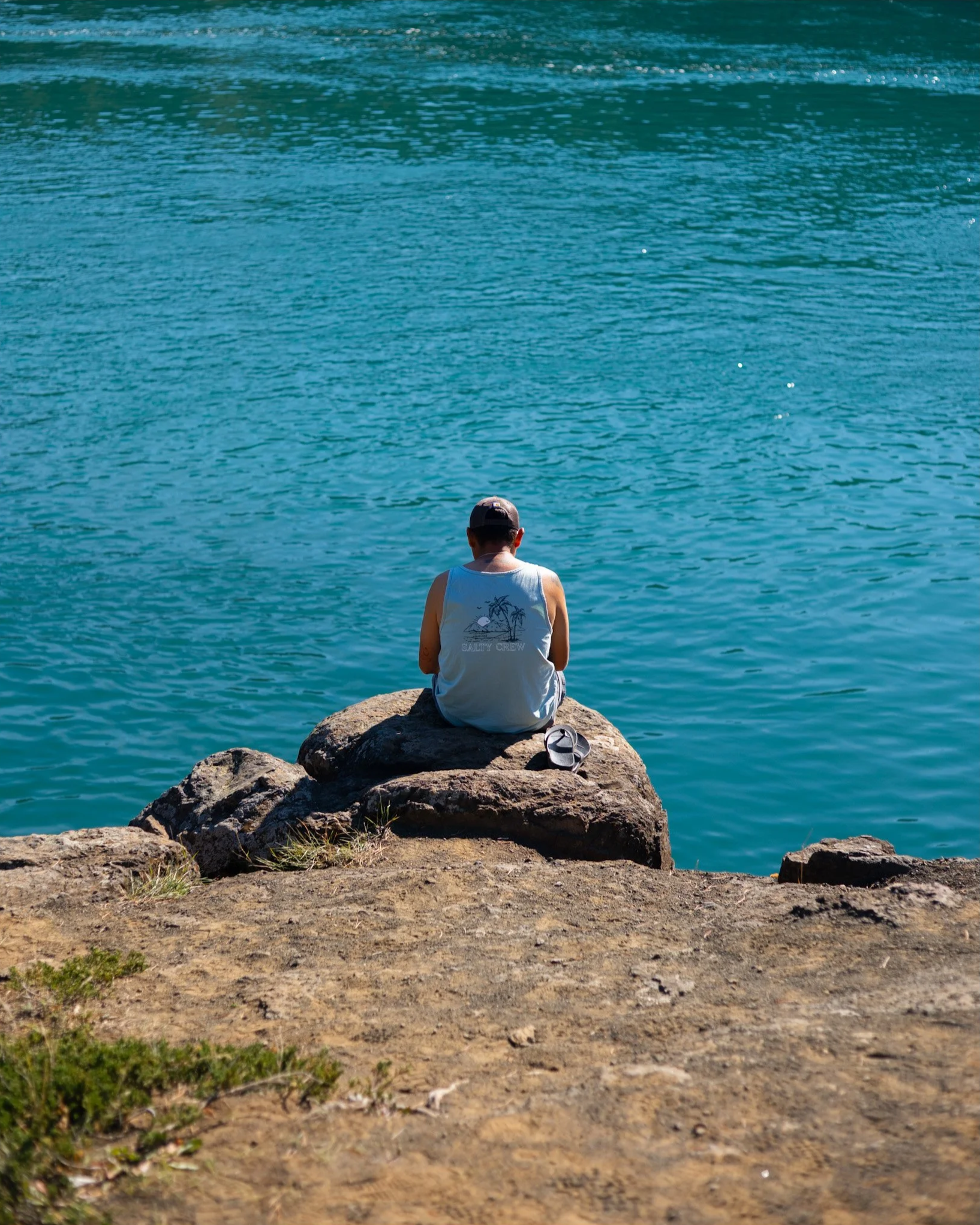 A man sitting on a large rock by a body of water, viewed from behind, wearing a tank top and a cap.