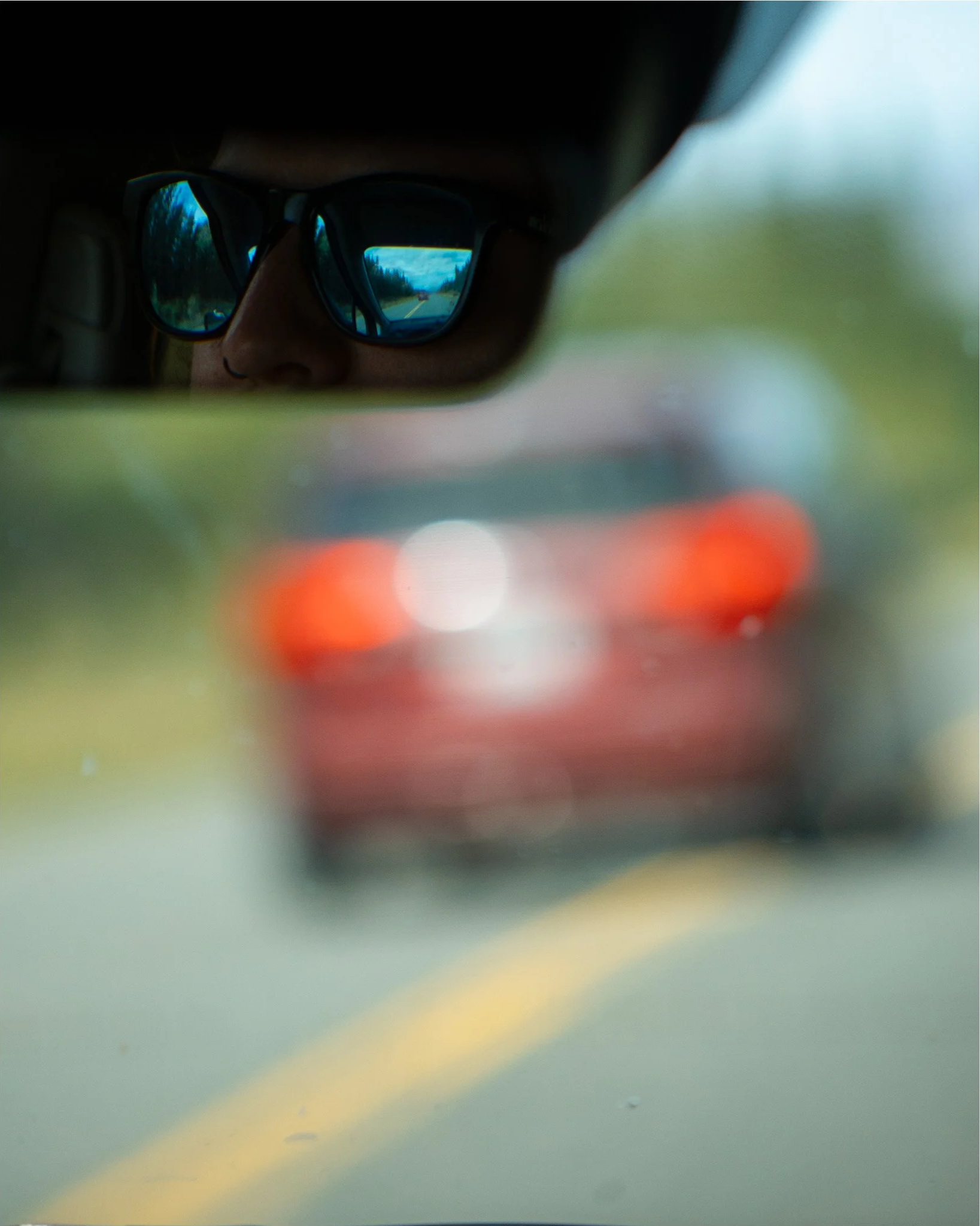 A person wearing sunglasses is reflected in the mirror, showing a view of a road with trees and a vehicle in the distance.