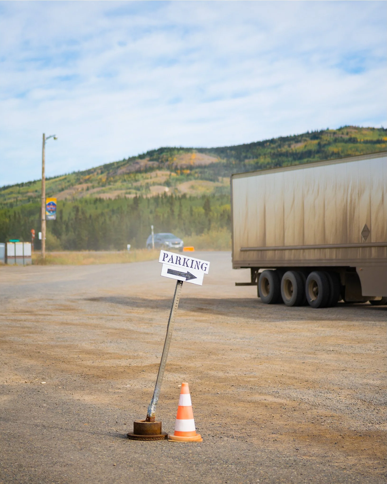 A parking lot with a small sign pointing to the right that says 'PARKING'. There is an orange and white traffic cone and a broken orange traffic cone next to a leaning, damaged pole. In the background, there are mountains, trees, a utility pole, and 