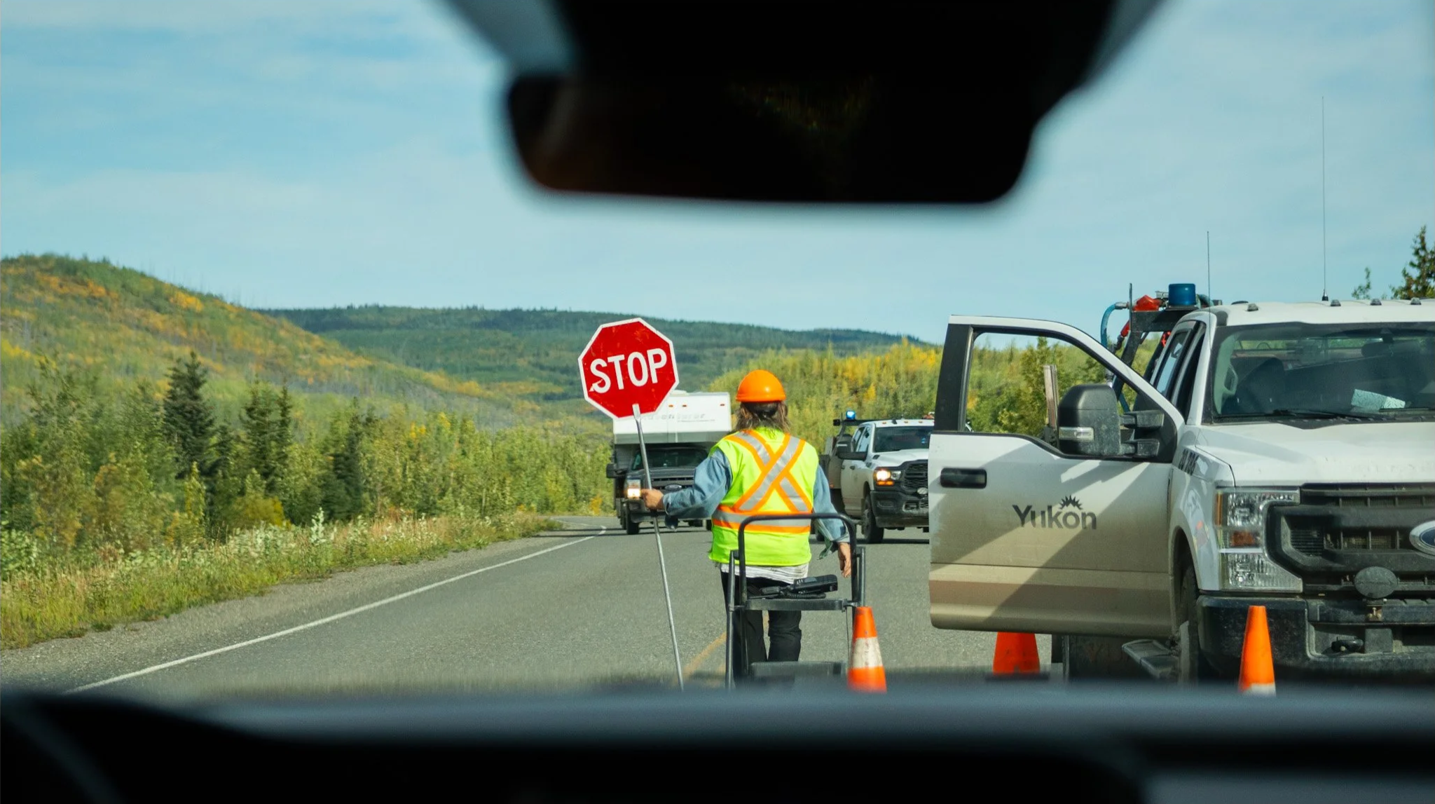 Highway scene with a traffic control worker in a blue vest and orange helmet, holding a stop sign, directing traffic. Several vehicles, including a Yukon truck and a police car, are stopped on a scenic road surrounded by green trees and rolling hills