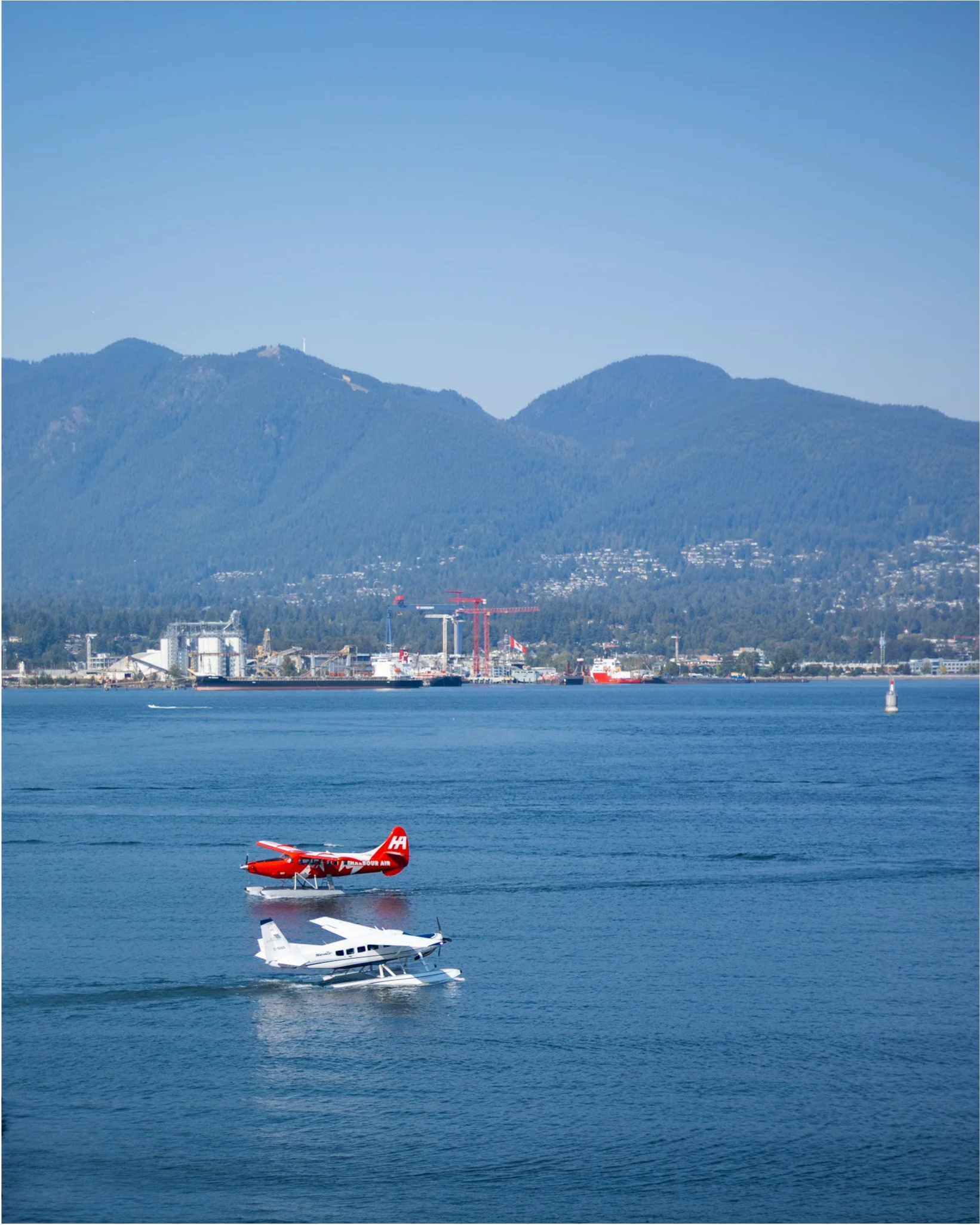 A seaplane taking off from the water with mountains in the background, a red floatplane in the foreground, and a large industrial structure near the shoreline.
