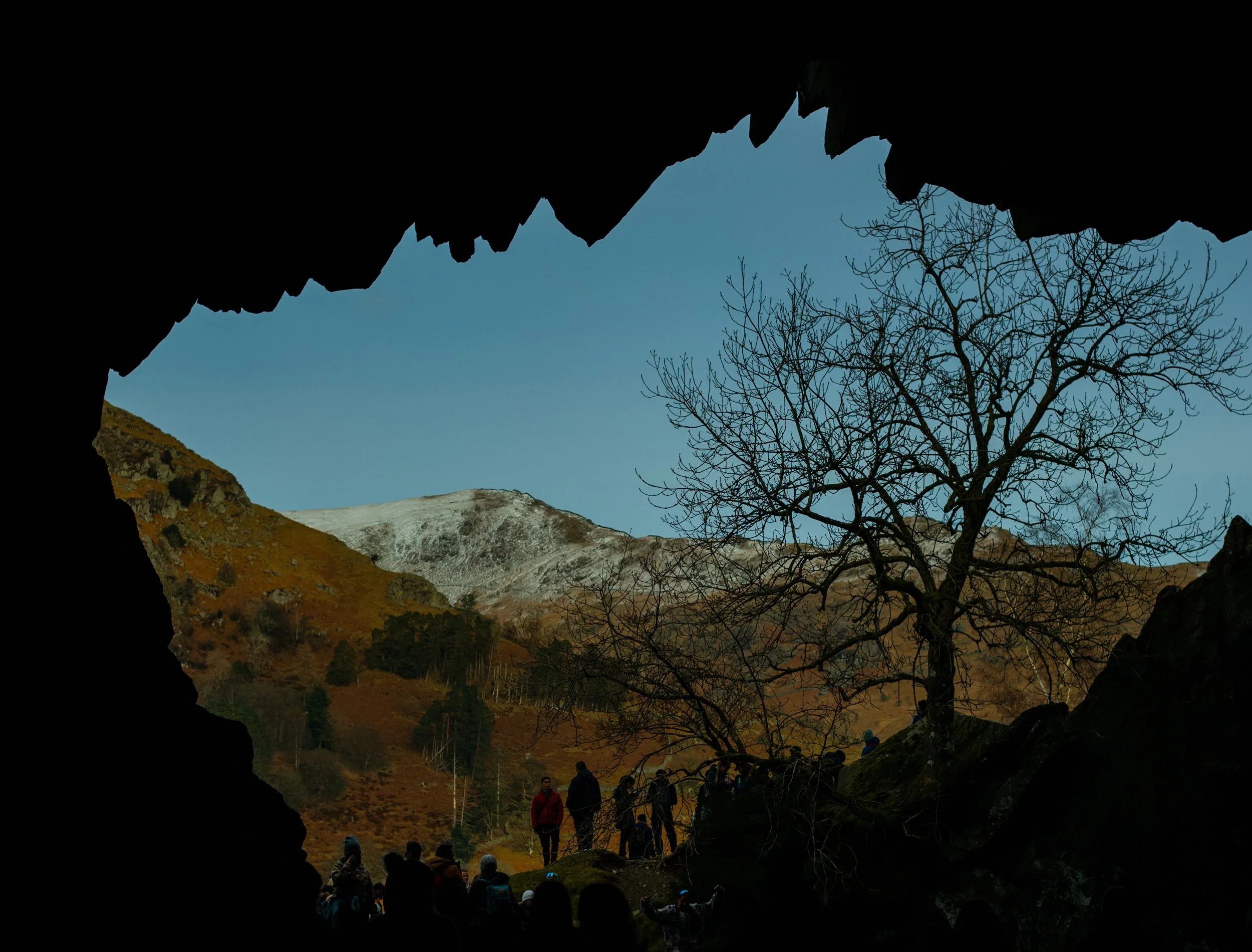 Group of hikers walking through a rocky mountain landscape with trees and snow-capped peaks in the distance, seen from inside a cave opening.
