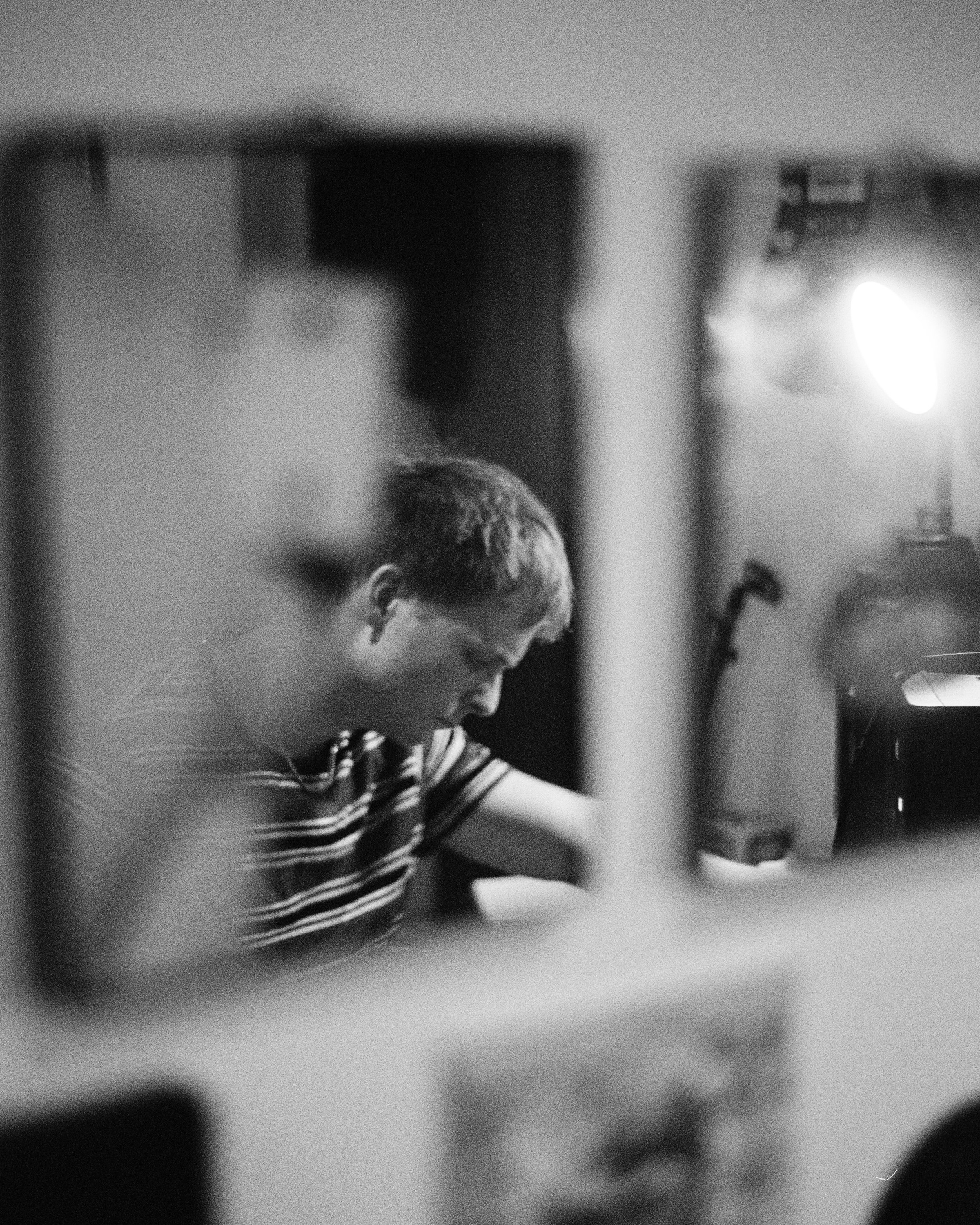 A young person with glasses and short hair, wearing a striped shirt, is looking downward while working on something on a table. There is a bright light source to the right side of the image, with a plain background.