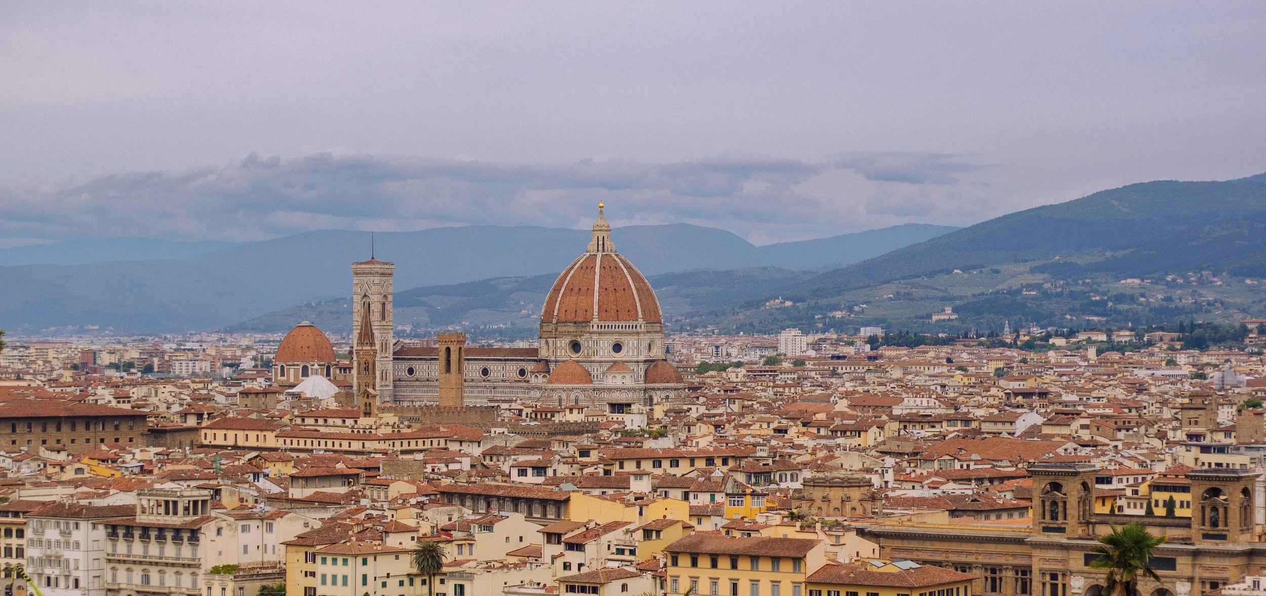 Landscape view of Florence, Italy, featuring the Cathedral of Santa Maria del Fiore (Duomo) with its red-tiled dome, surrounded by historic buildings and mountains in the background.