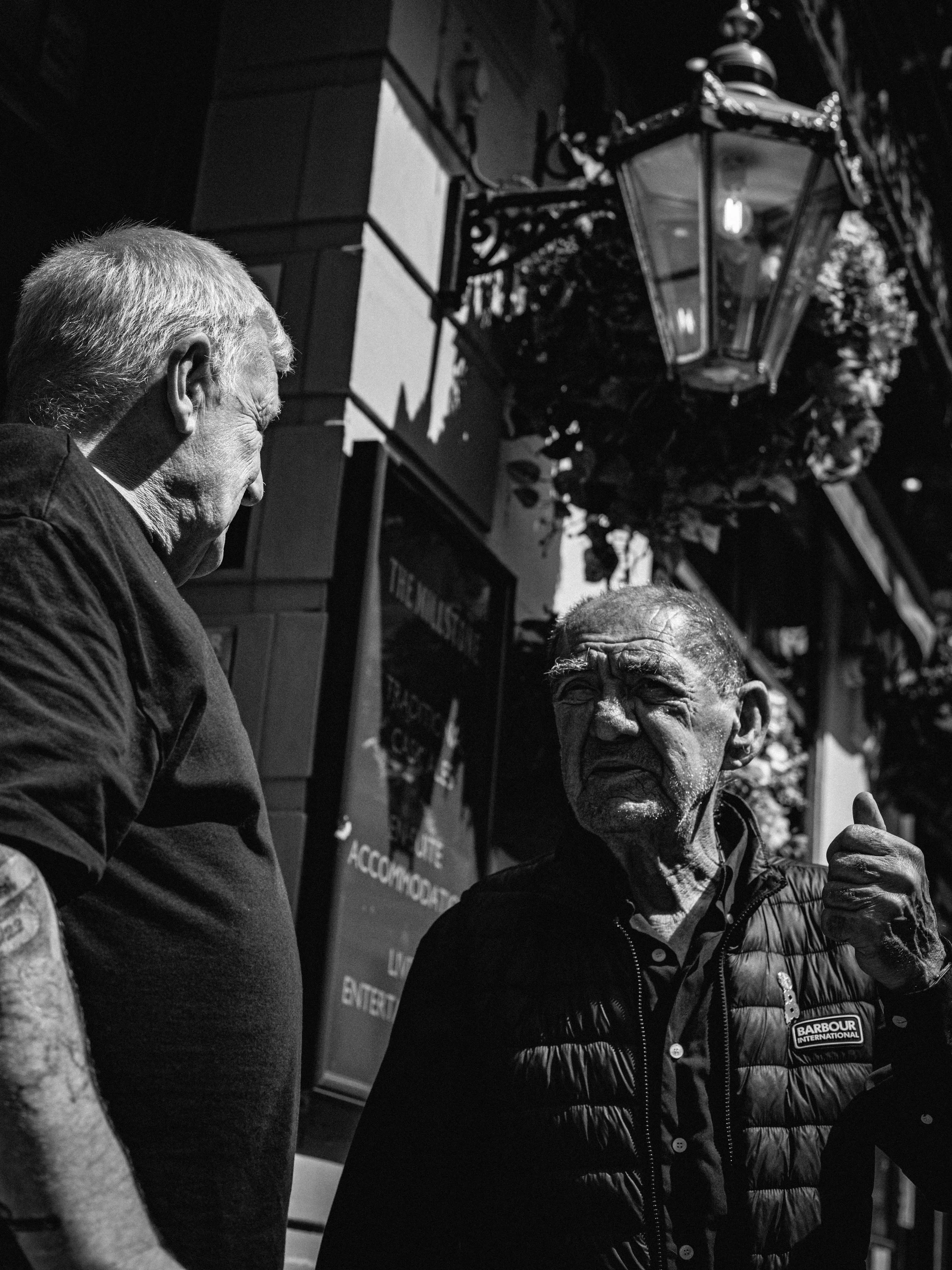 Two older men are engaged in conversation outside during nighttime. One man on the left has gray hair and is looking downward, while the man on the right is wearing glasses and a black puffy jacket with a 'Barbour International' logo, and is pointing