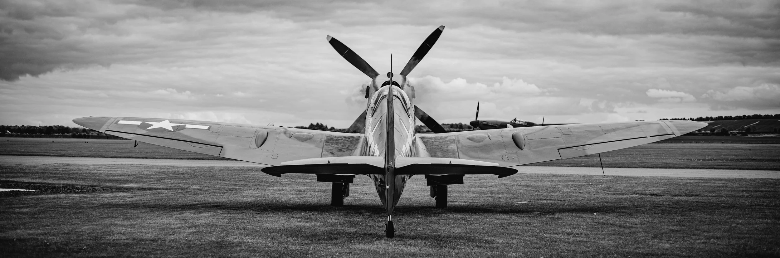 Black and white photo of a vintage military aircraft taxiing on a runway, with another aircraft in the background under cloudy skies.