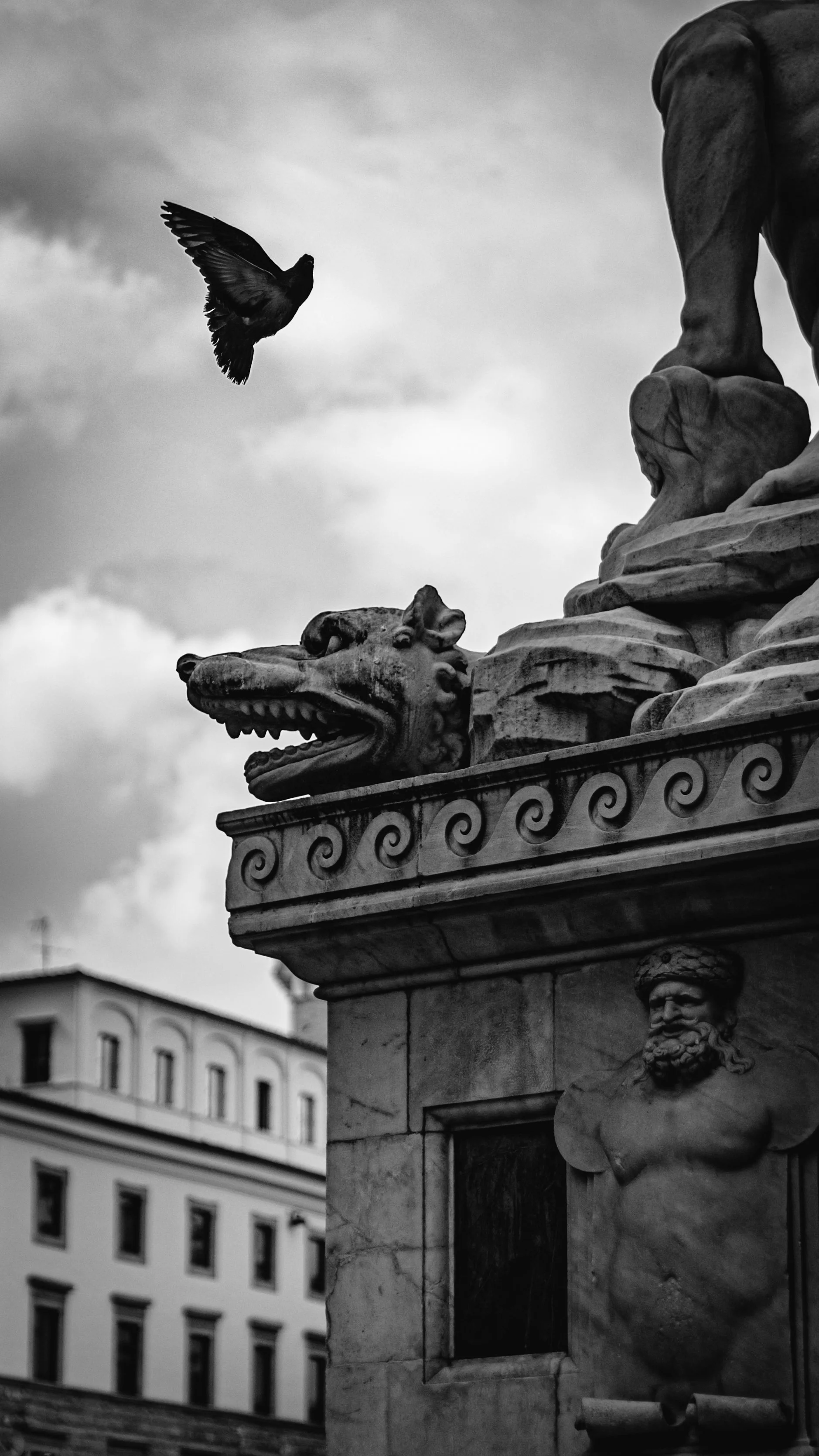 Black and white photo of a decorative stone fountain with a lion's head sculpture and a bearded man, with a bird flying nearby in front of a cloudy sky and city buildings in the background.