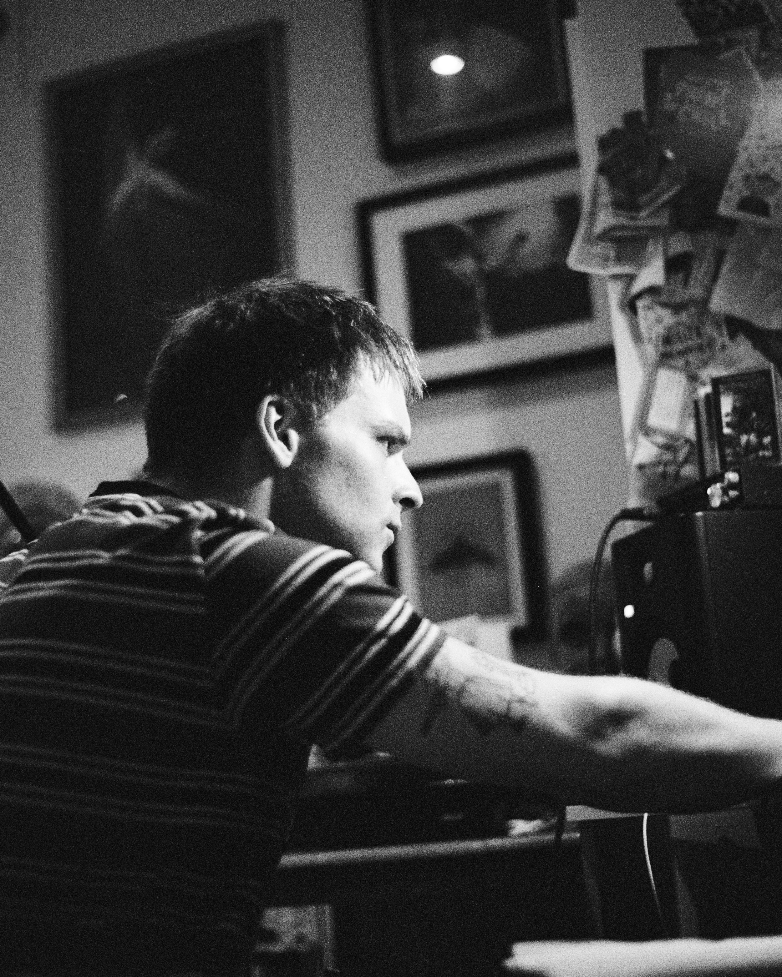 A person with short dark hair wearing a striped shirt, sitting and working at a computer with a monitor and various papers around, in a black and white photo.